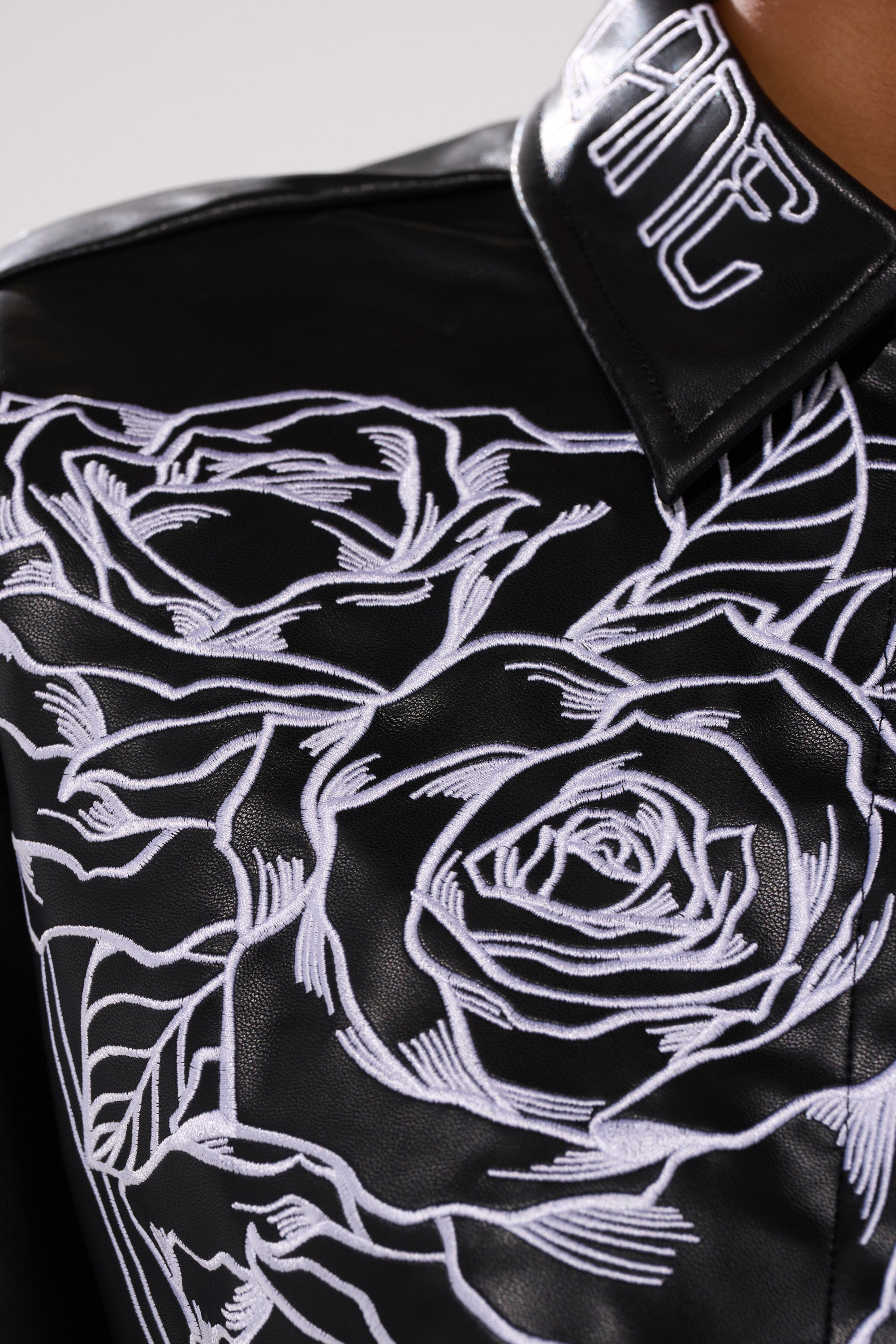 Close-up of the MAKE IT OFFICIAL FAUX LEATHER BUTTON DOWN IN BLACK, showcasing white floral embroidery with large roses and abstract lines, plus embroidered white lettering on the collar.