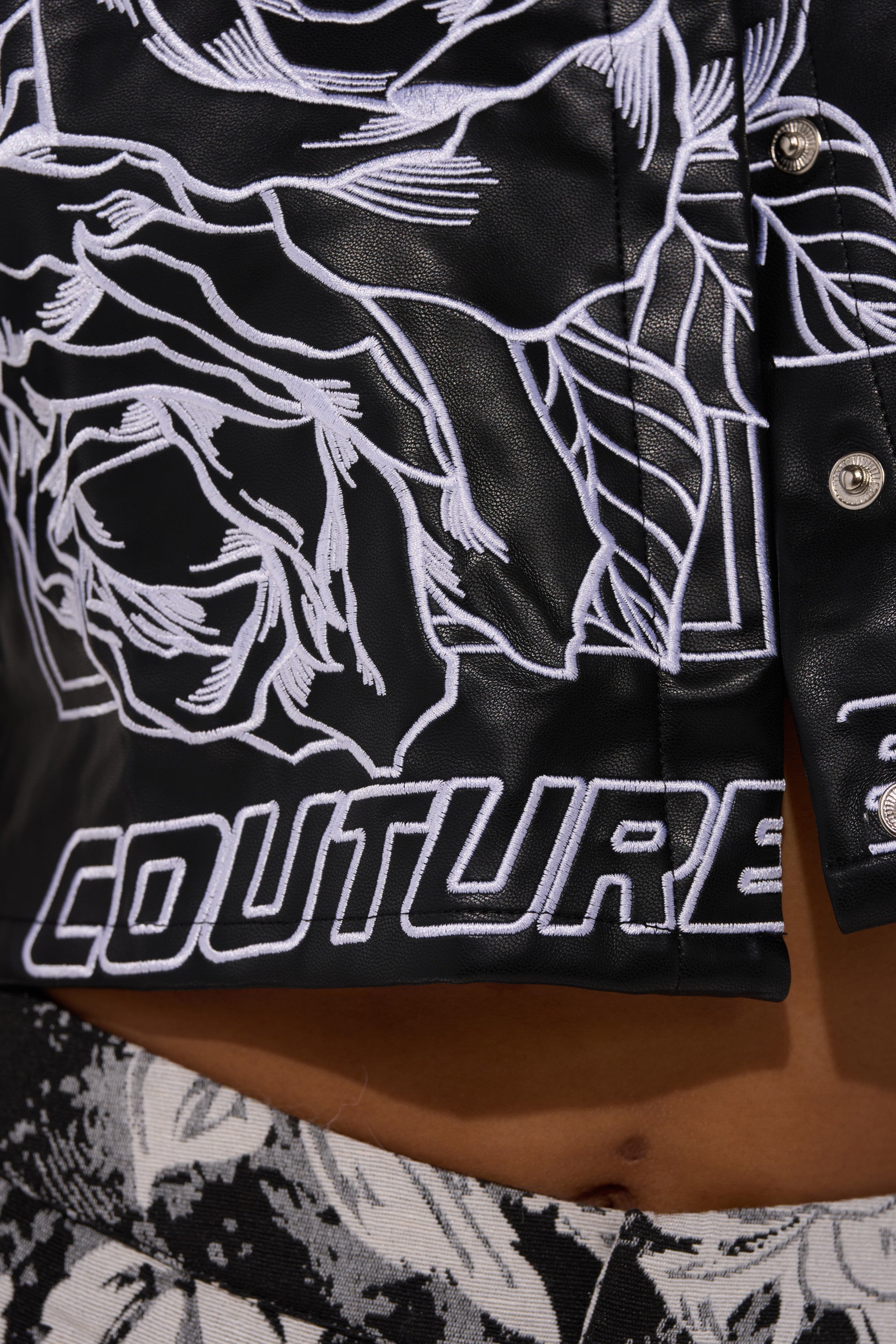 Close-up of the MAKE IT OFFICIAL FAUX LEATHER BUTTON DOWN IN BLACK, featuring white floral stitching and "COUTURE" embroidery near the hem, worn over patterned pants with a hint of bare midriff visible.