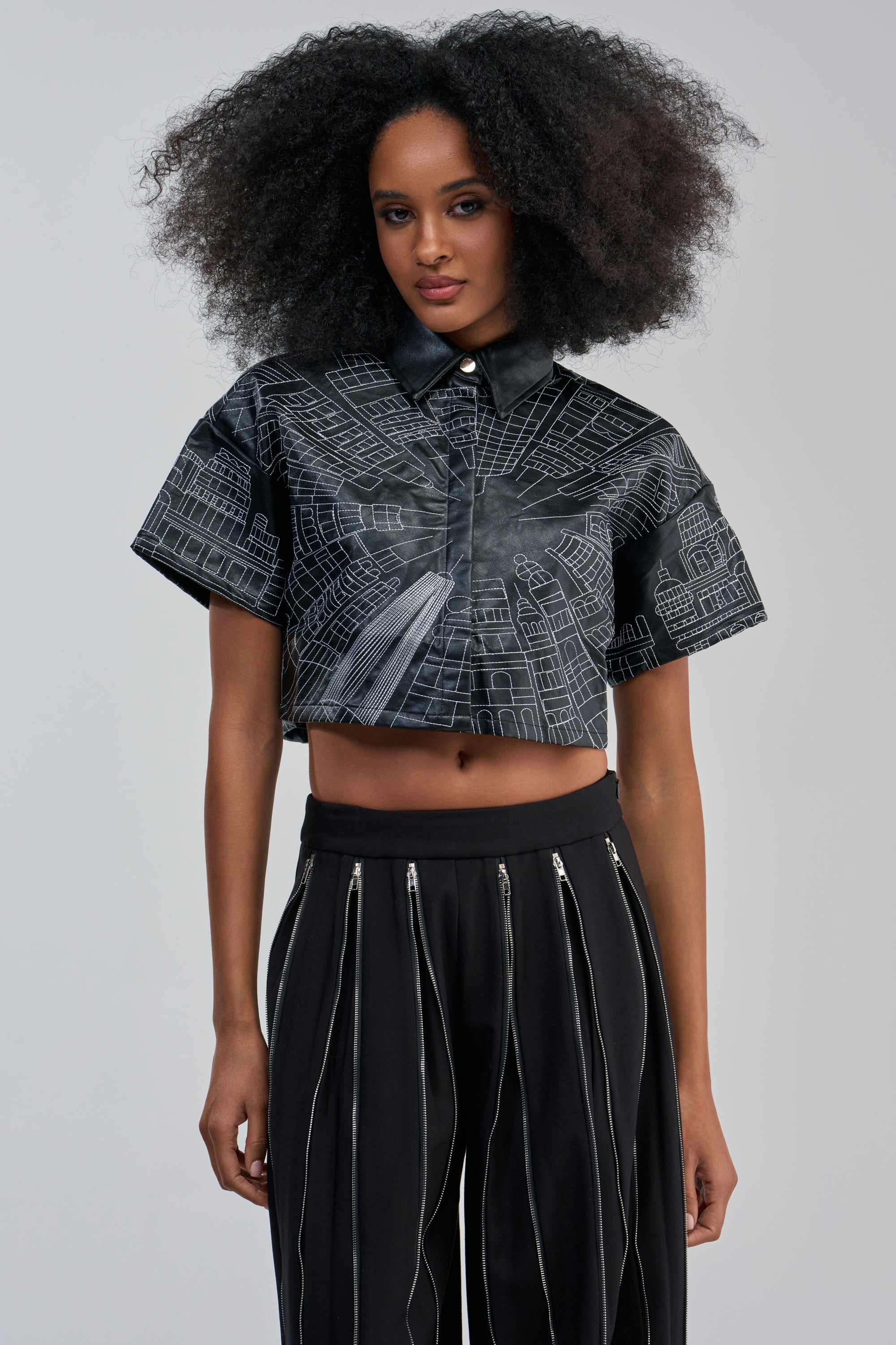 A woman with voluminous curly hair wears the DONT LOOK UP PU CROPPED BUTTON DOWN— a black cropped shirt with skyline embroidery—paired with high-waisted black pants featuring vertical zippers for a striking streetwear look. She stands against a plain light background.