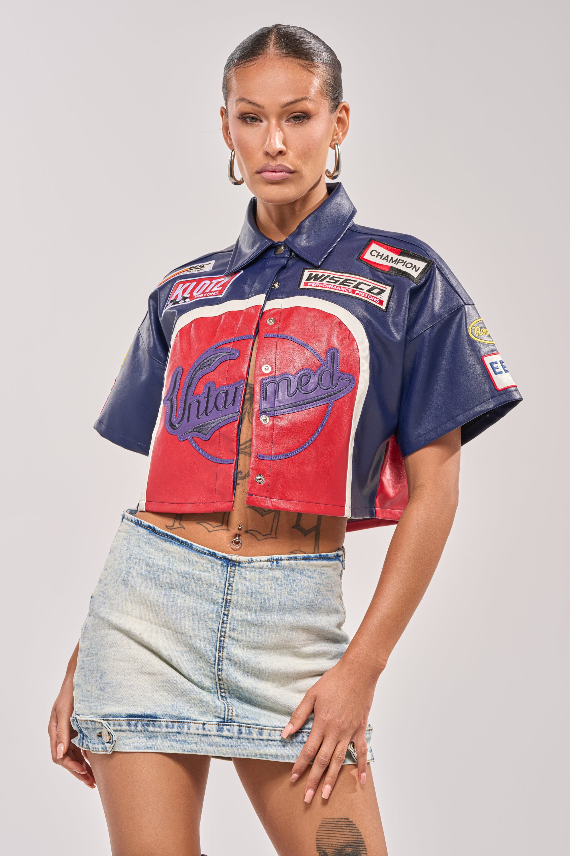 Wearing the PEDAL TO THE METAL FAUX LEATHER BUTTON DOWN, hoop earrings, and a light blue denim mini skirt, a woman channels bold streetwear style as she stands confidently against a plain light background. 1