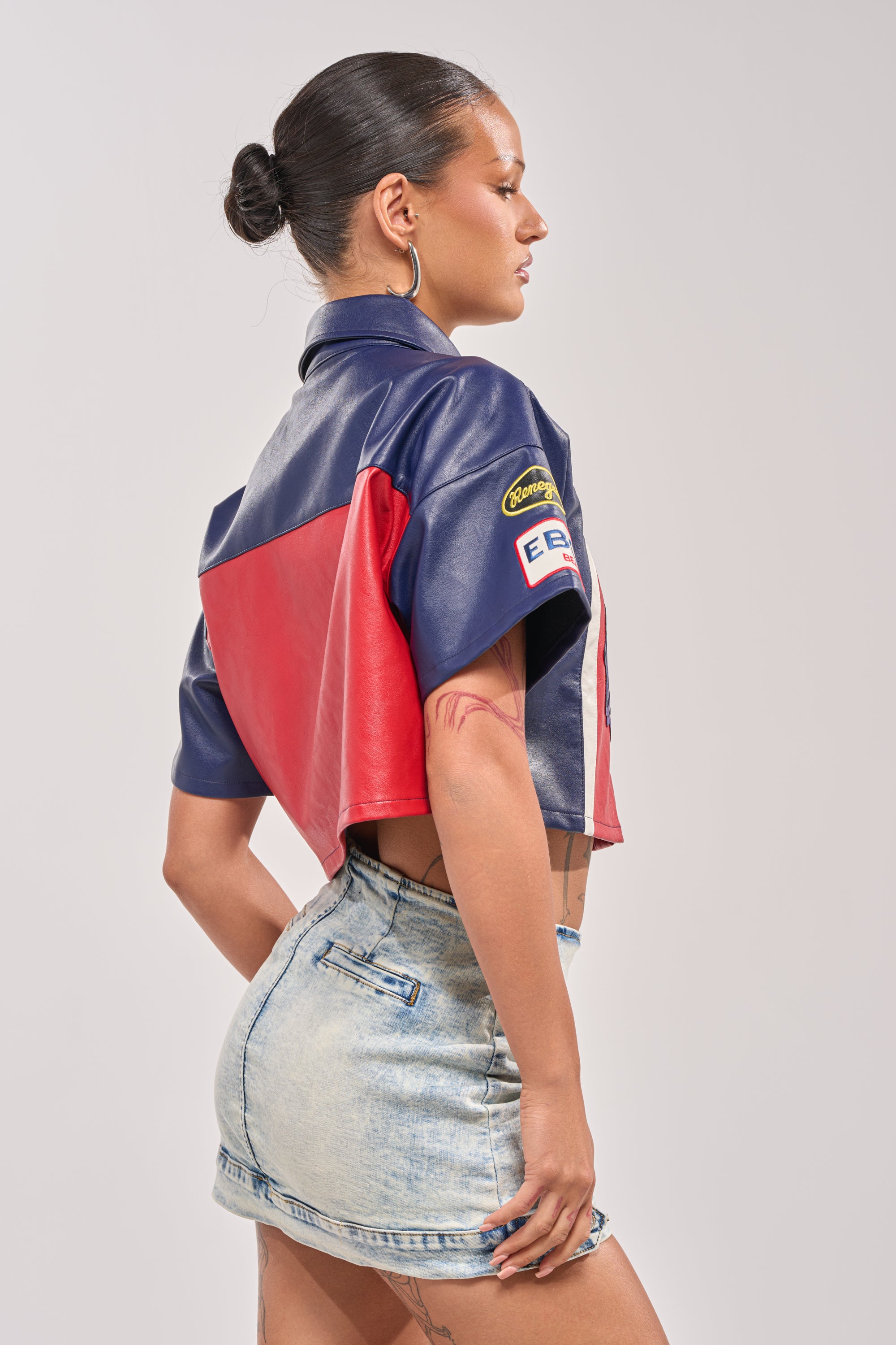 A woman in profile wears the PEDAL TO THE METAL FAUX LEATHER BUTTON DOWN with patches, paired with light-wash denim shorts. She styles her hair in a sleek bun, sports hoop earrings and shows off tattoos for a cool streetwear look. 2