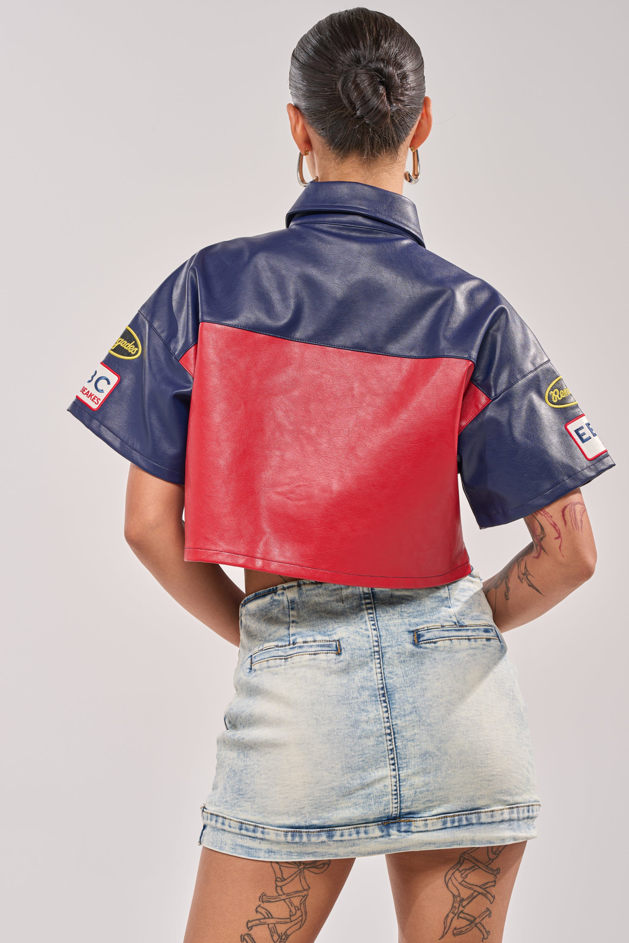 A person with hair in a bun is seen from behind in streetwear, featuring the PEDAL TO THE METAL FAUX LEATHER BUTTON DOWN, a cropped red and navy jacket with sleeve patches, paired with a high-waisted light denim mini skirt. Tattoos are visible on their legs. 3