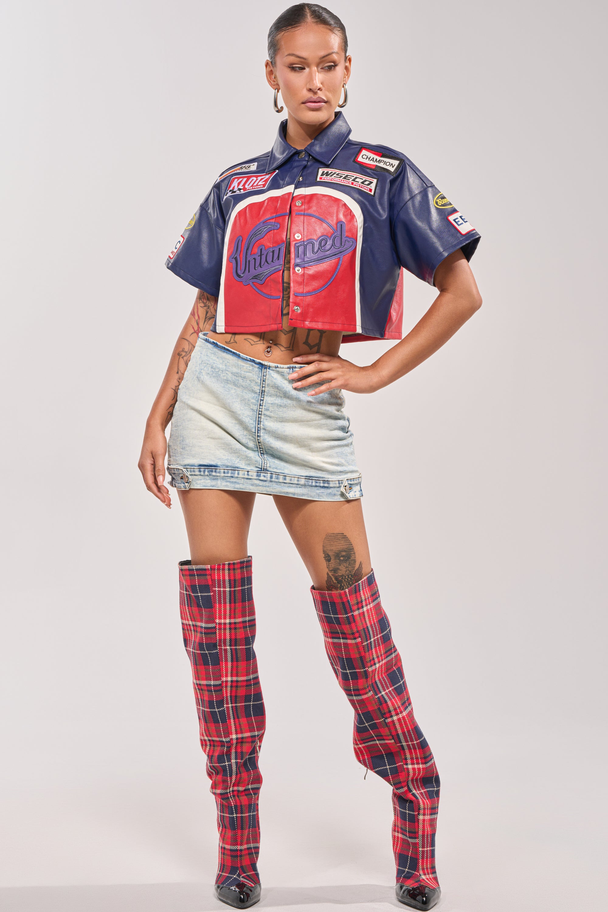 A woman poses confidently in streetwear, wearing the PEDAL TO THE METAL FAUX LEATHER BUTTON DOWN, a light denim mini skirt, tall red plaid boots, and hoop earrings. Her tattoos are visible as she stands against a plain light background. 4