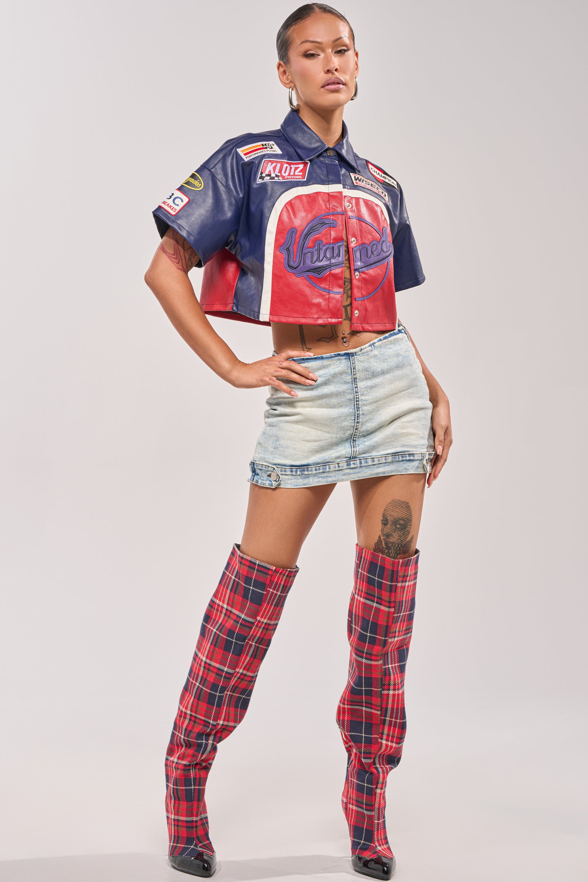 A woman rocks the PEDAL TO THE METAL FAUX LEATHER BUTTON DOWN with a light denim mini skirt and red plaid thigh-high boots, pairing hoop earrings and slicked-back hair for bold festival-ready street style. 5