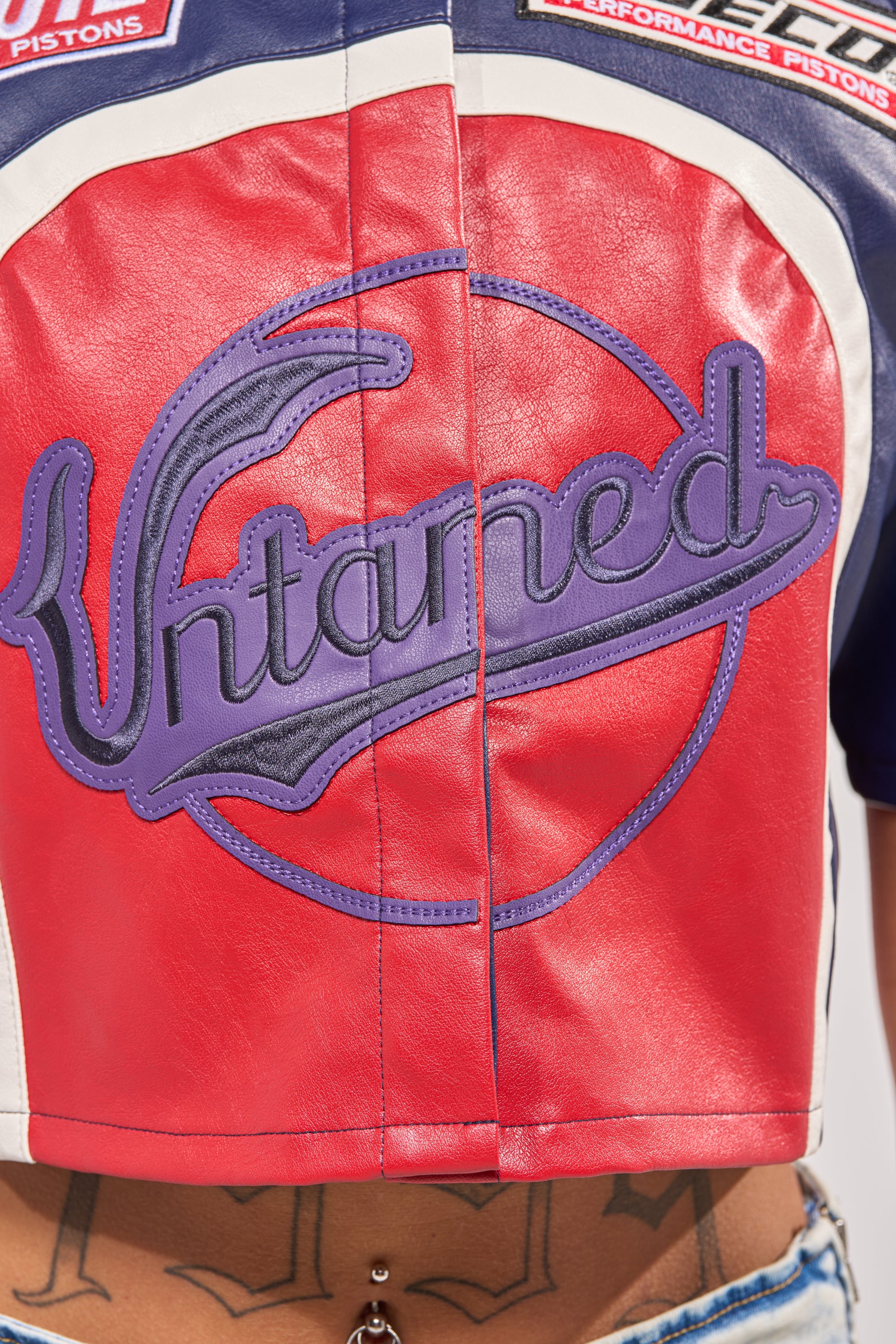 Close-up of the PEDAL TO THE METAL FAUX LEATHER BUTTON DOWN, a red and blue racing-inspired piece with bold purple "Untamed" embroidery. Tattoo and belly button piercing peek out—ideal for daring streetwear looks. 6