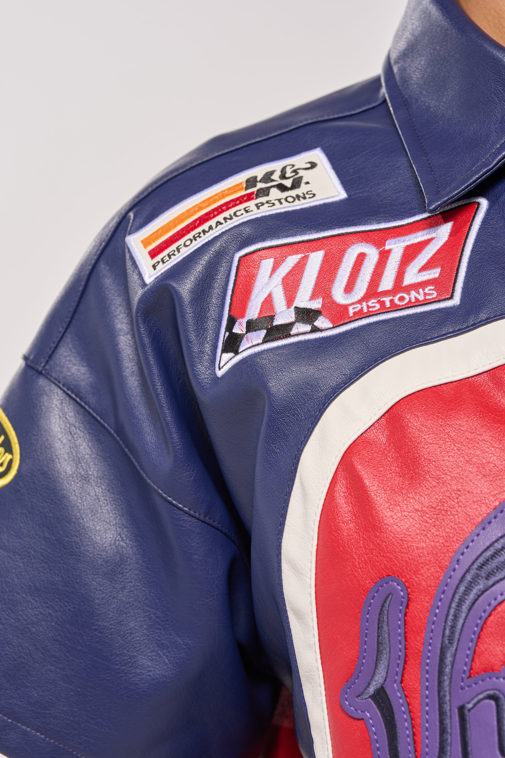 Close-up of the PEDAL TO THE METAL FAUX LEATHER BUTTON DOWN, a blue and red jacket with racing-themed patches like "KLOTZ Pistons," a checkered flag, and "Performance Pistons"—a standout piece for bold streetwear looks. 7