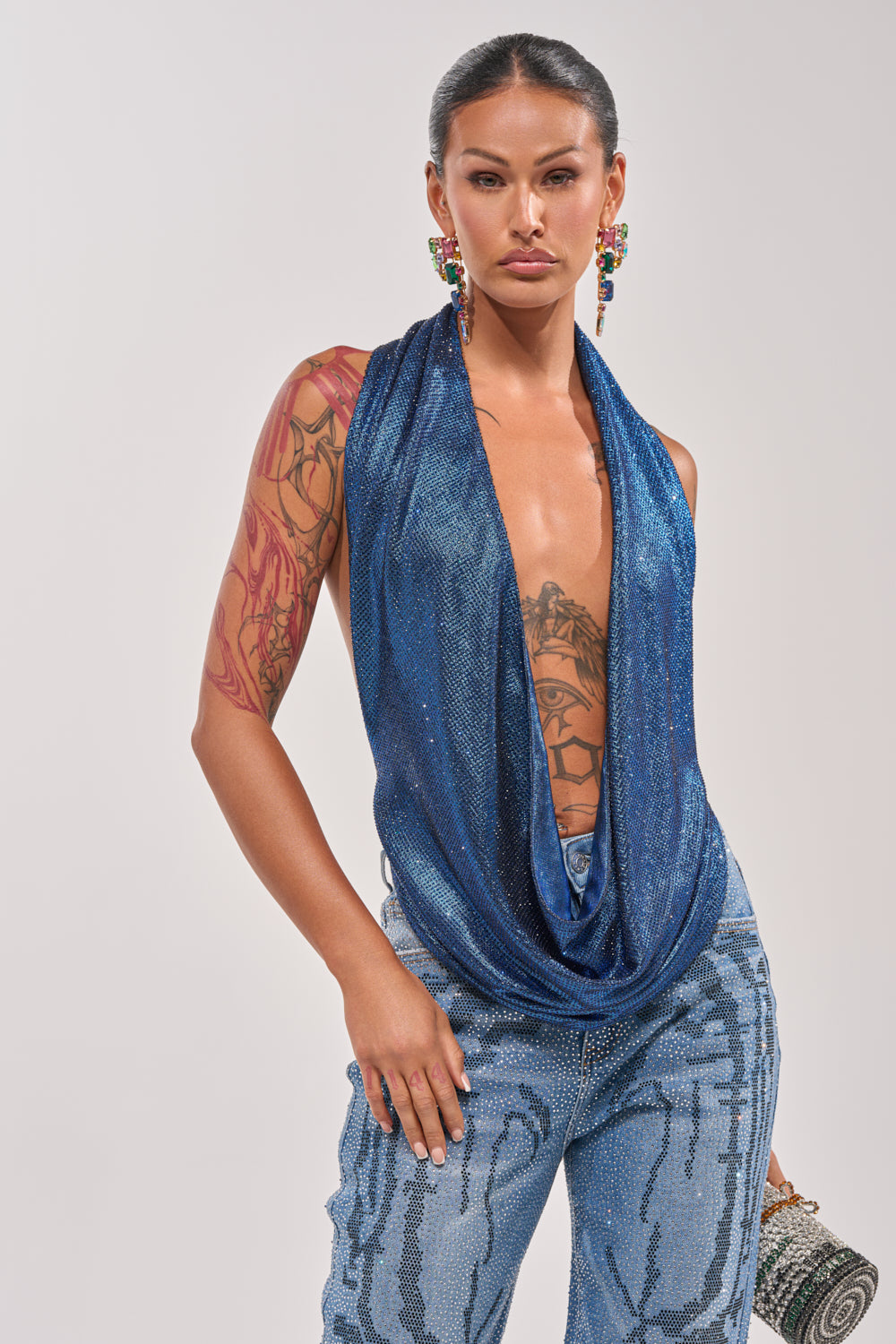 A woman with long dark hair in a sleek ponytail wears the MERMAID DREAM RHINESTONE OPEN BACK HALTER TOP, sequined blue jeans, colorful beaded earrings, visible arm tattoos, and holds a small textured clutch. 1