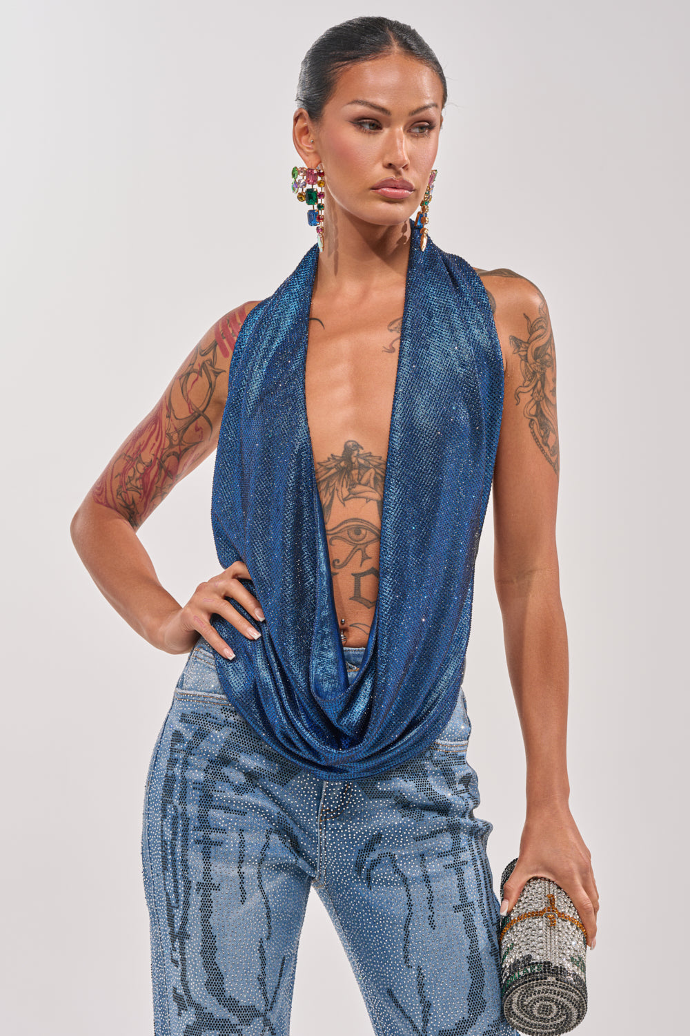 A woman with tattooed arms wears the MERMAID DREAM RHINESTONE OPEN BACK HALTER TOP and patterned blue pants, holding a beaded clutch and wearing colorful earrings as she stands confidently against a plain background. 2