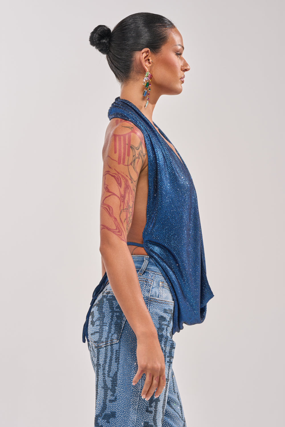 A woman with dark hair in a bun, colorful earrings, and an arm tattoo poses in profile wearing the MERMAID DREAM RHINESTONE OPEN BACK HALTER TOP and light blue patterned jeans against a plain background. 3