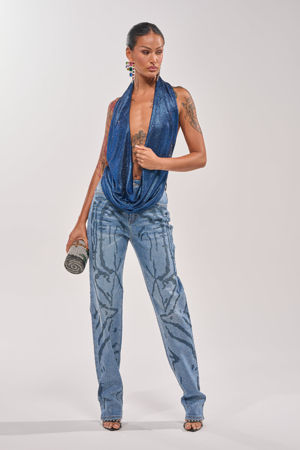 A woman poses confidently in the MERMAID DREAM RHINESTONE OPEN BACK HALTER TOP and shimmering light blue sequined pants with wavy patterns, accessorized with a black and silver clutch, open-toe heels, and showcasing her arm tattoos. 5