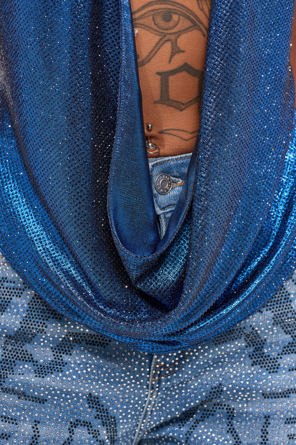 Wearing the MERMAID DREAM RHINESTONE OPEN BACK HALTER TOP and sparkly jeans, a person reveals a lower back tattoo of an eye and geometric shapes, along with a belly button piercing. 8