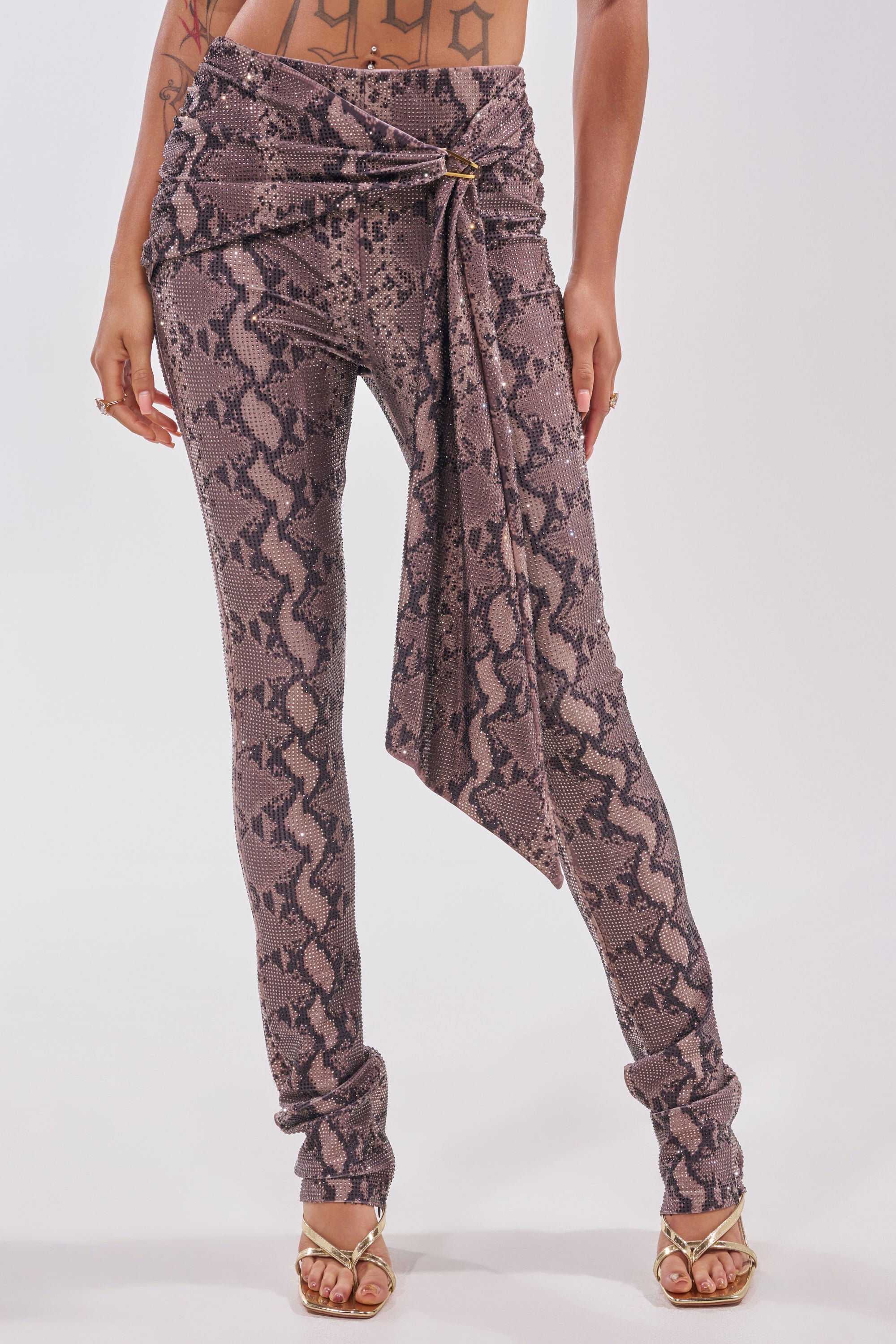 A person wearing the VENOM KISS RHINESTONE LEGGING—high-waisted brown and black snake-print flares with a draped sash at the waist—paired with gold high-heeled sandals. Only the lower half of their body is shown. 1