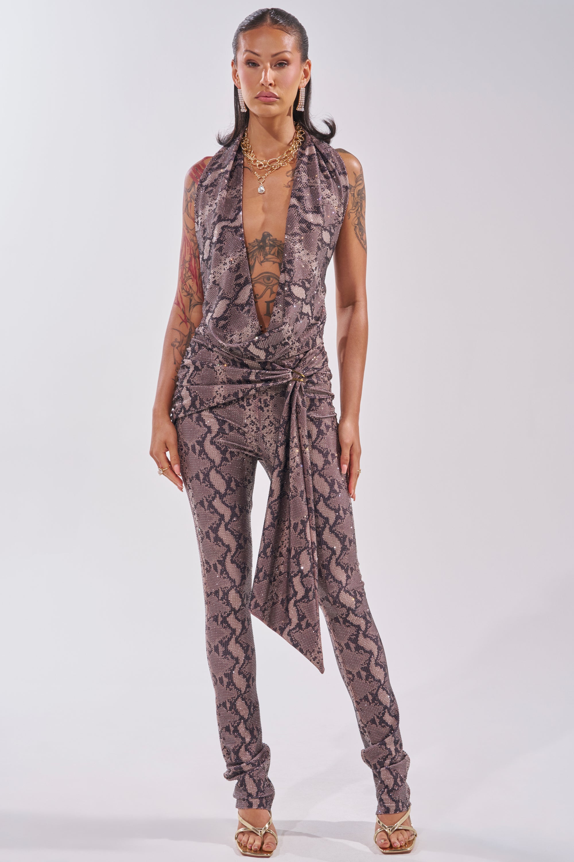 A woman stands confidently in the VENOM KISS RHINESTONE HALTER TOP with a deep V-neck and snake print, paired with gold jewelry and strappy heels. She displays visible arm tattoos against a plain, light-colored background. 6