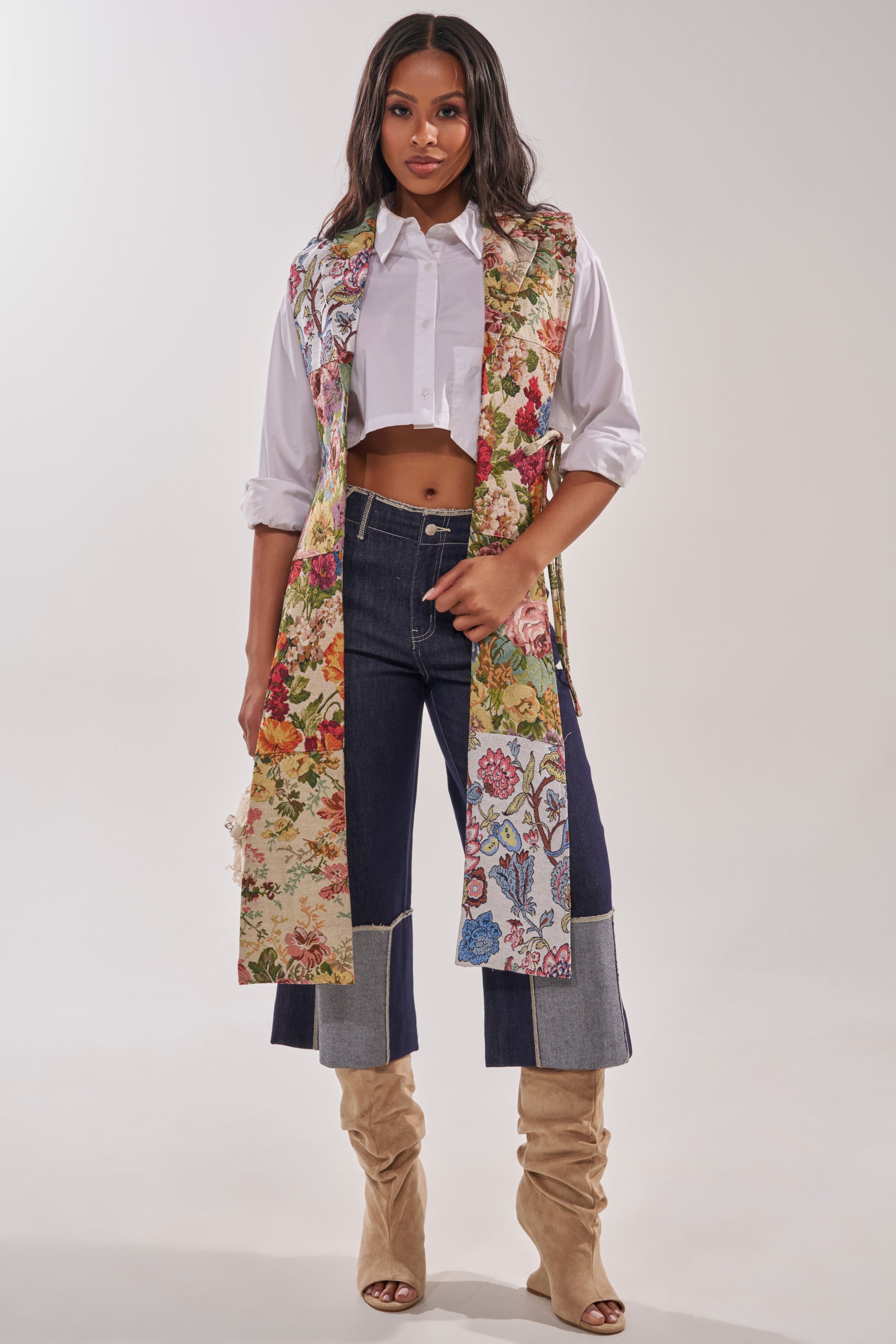 A woman stands confidently against a plain background wearing the HONEY HONEY SPLIT VEST IN TAPESTRY over a white cropped shirt, high-waisted dark cuffed jeans, and tan knee-high boots. 0