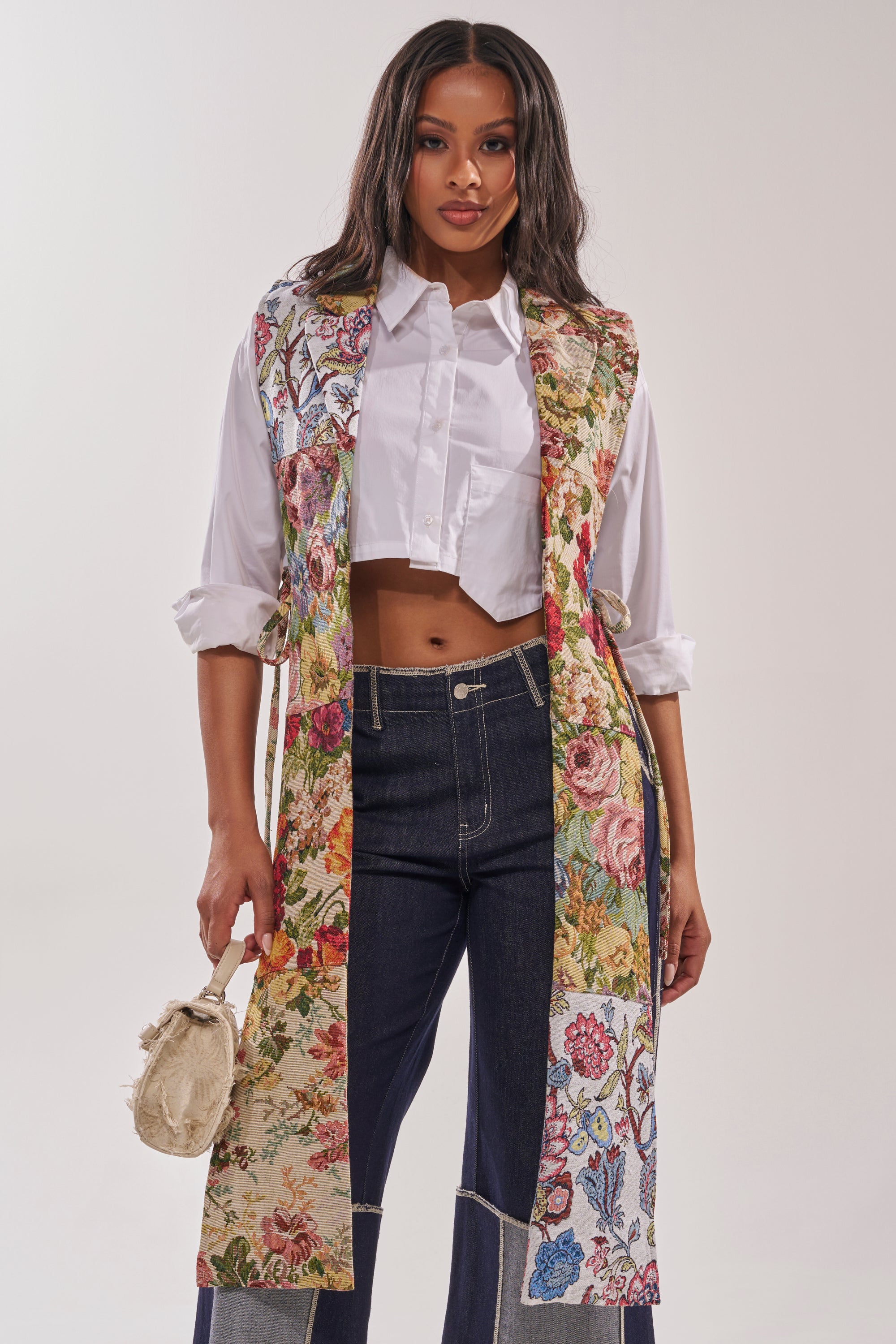 A woman stands facing forward against a plain light background, wearing the HONEY HONEY SPLIT VEST IN TAPESTRY over a white cropped shirt and dark high-waisted jeans, holding a small beige purse. 1