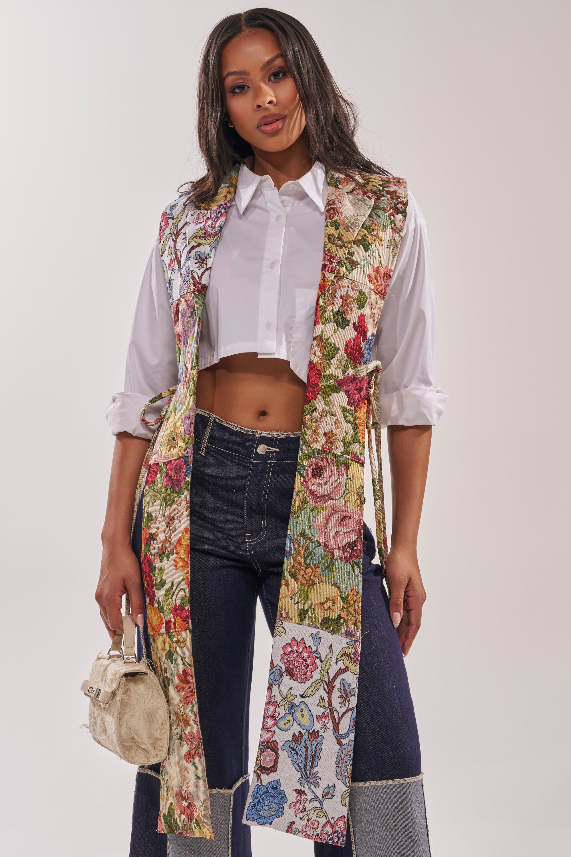 A woman models the HONEY HONEY SPLIT VEST IN TAPESTRY over a white cropped button-up and high-waisted two-tone jeans, holding a small beige handbag against a simple light background. 2