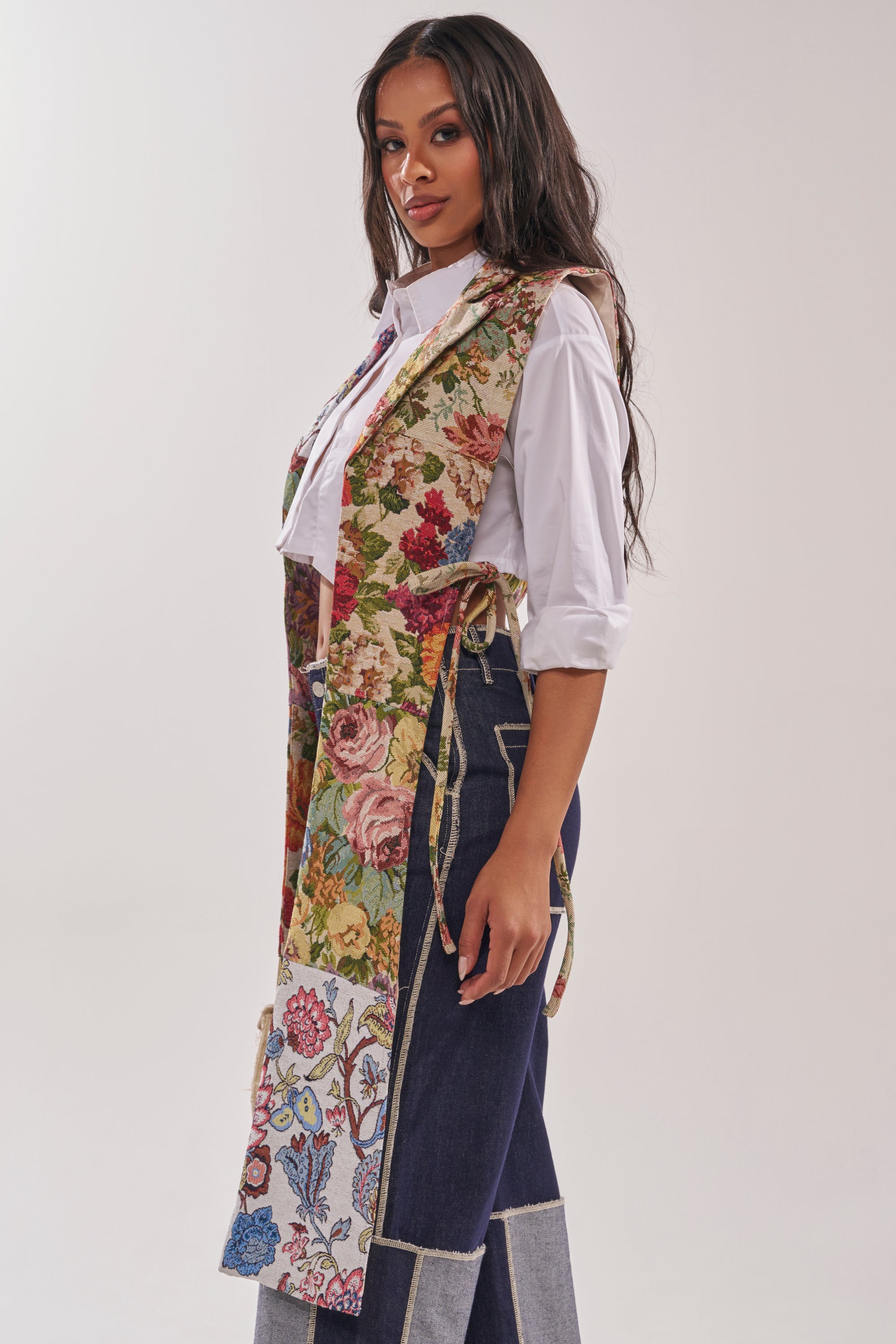 A woman stands sideways against a plain light background, wearing a white shirt, high-waisted blue jeans with patchwork details, and the HONEY HONEY SPLIT VEST IN TAPESTRY featuring side ties. 3