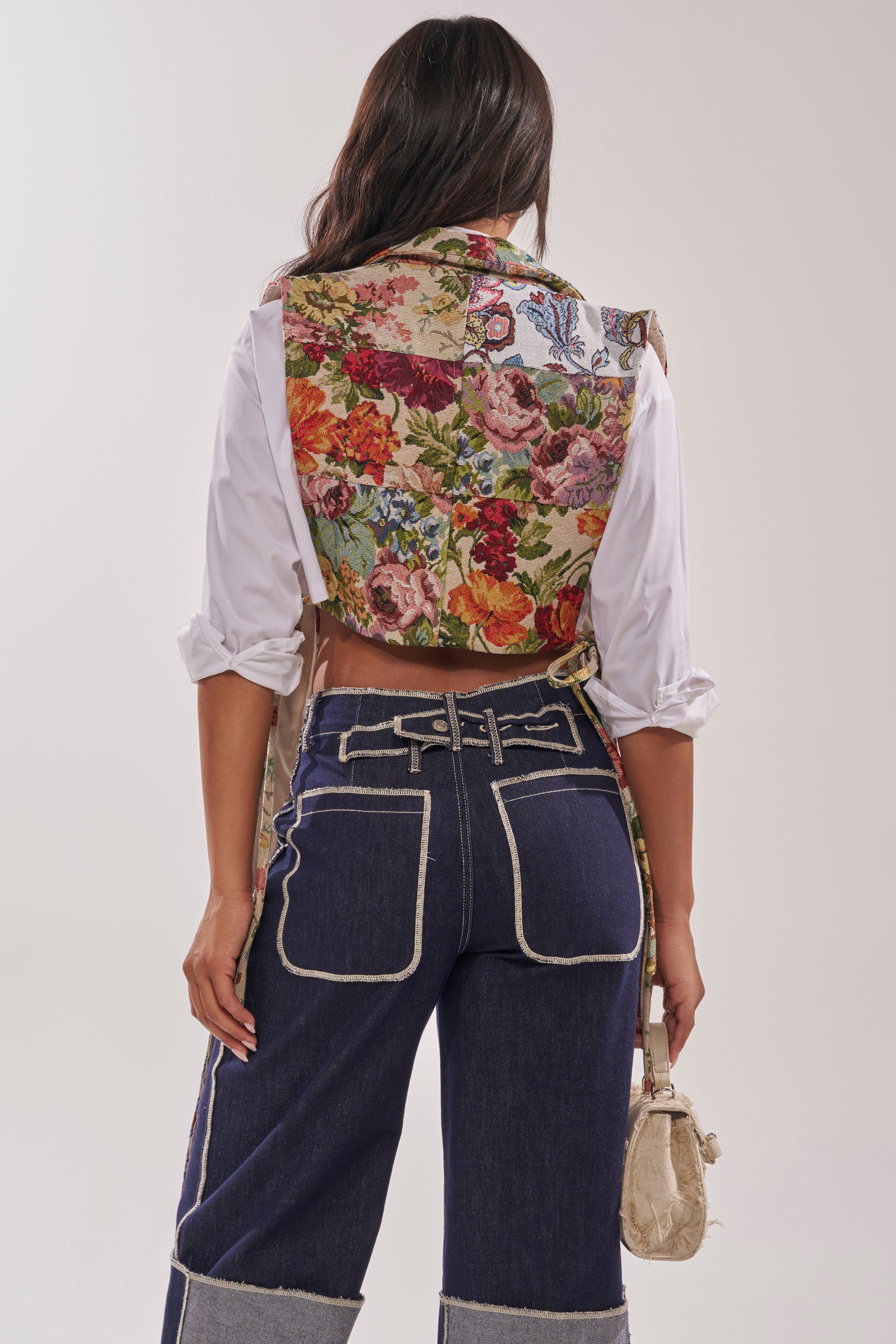A person stands facing away, wearing the HONEY HONEY SPLIT VEST IN TAPESTRY over a rolled-sleeve white shirt and dark denim jeans with white stitching and cuffed hems, holding a small beige bag. 4