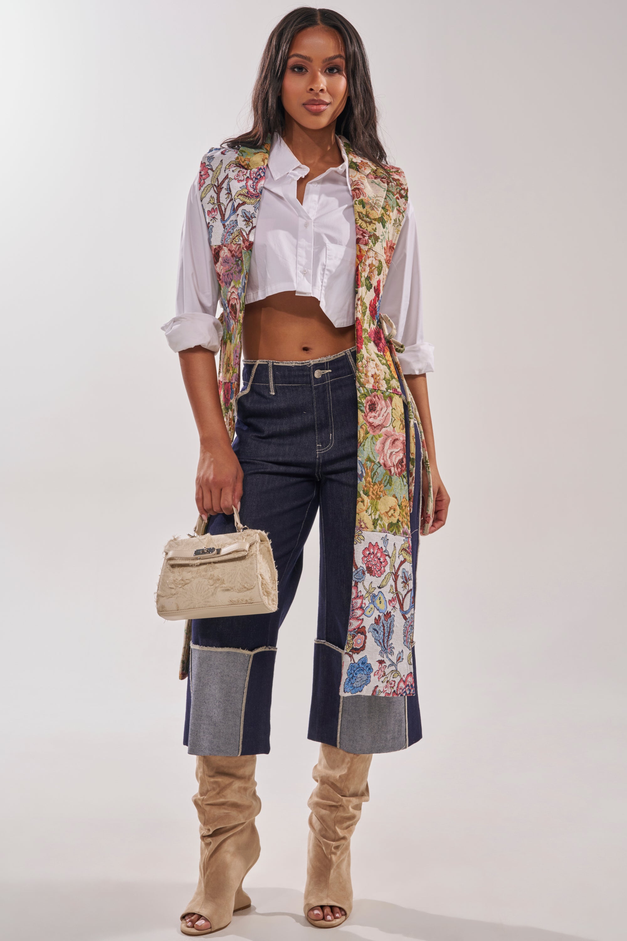A woman poses against a plain light background, wearing the HONEY HONEY SPLIT VEST IN TAPESTRY over a white cropped shirt, dark wide-leg jeans with light cuffs, beige knee-high boots, and holding a small beige handbag. 5
