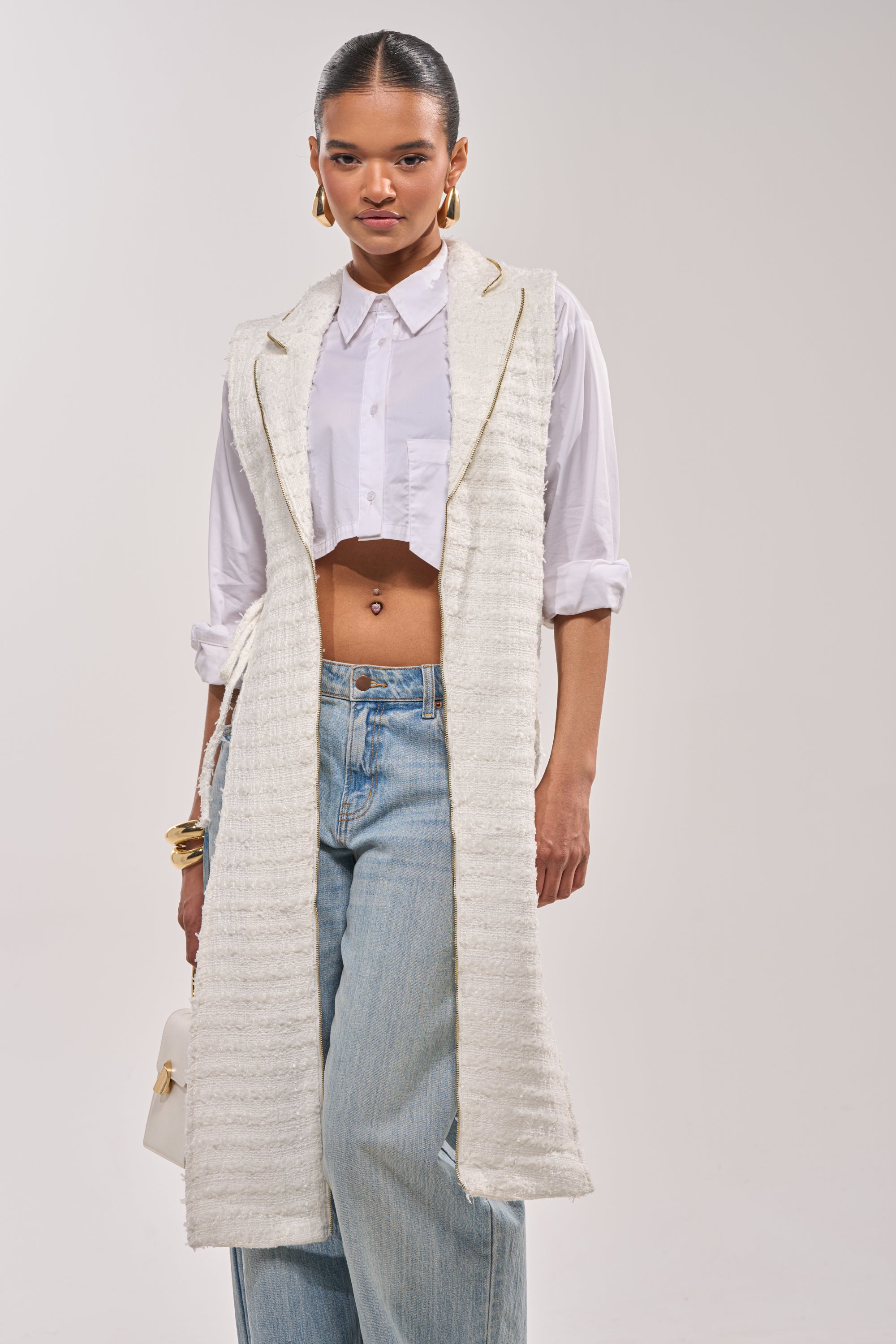 A woman wears a white cropped button-up, light blue wide-leg jeans, and the HONEY HONEY SPLIT VEST IN TWEED. She accessorizes with gold jewelry, carries a small white handbag, and stands against a neutral background. 0