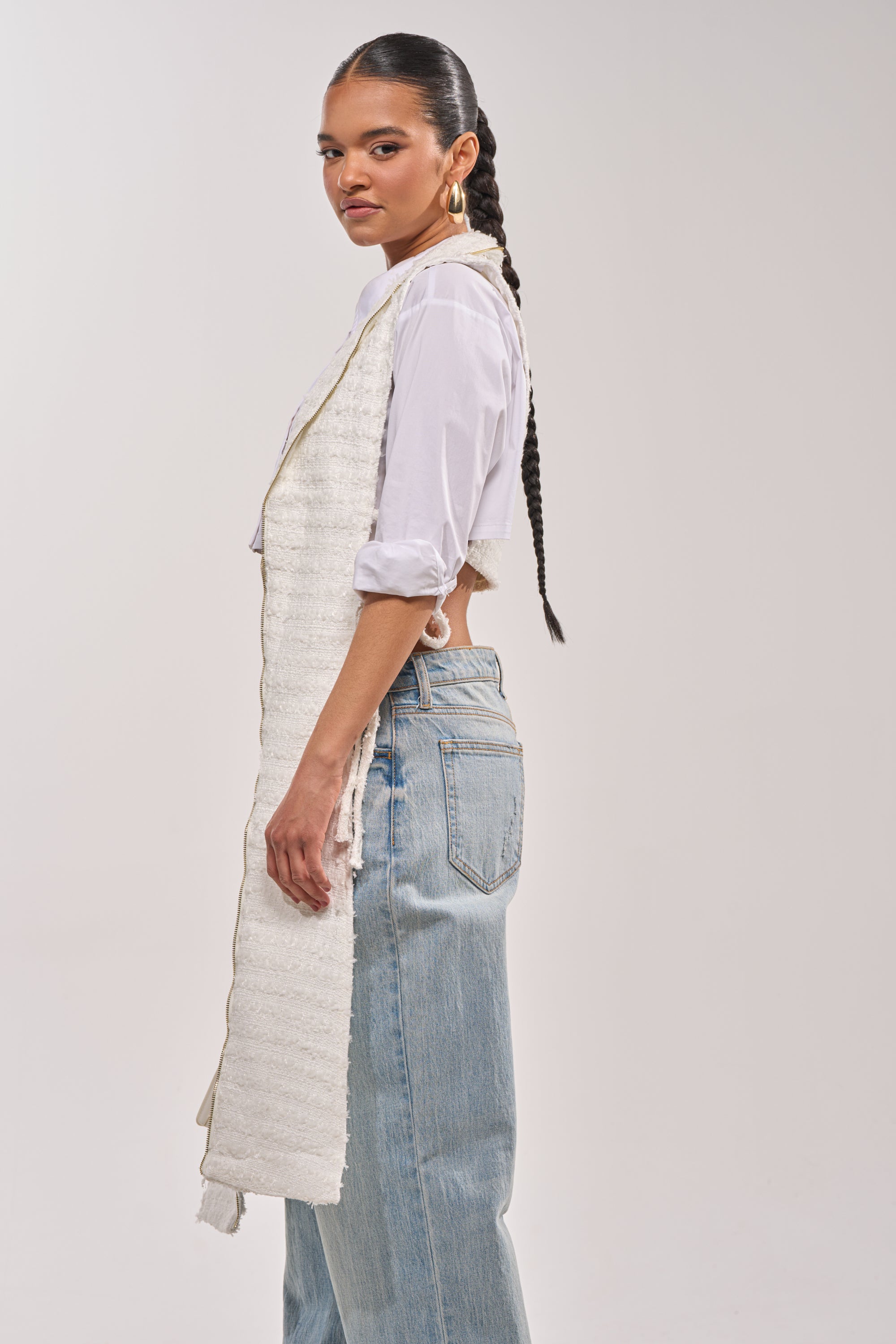 A woman with a long braided ponytail pairs a white shirt and light blue high-waisted jeans with the HONEY HONEY SPLIT VEST IN TWEED, posing against a plain light background for an effortlessly modern night-out look. 1