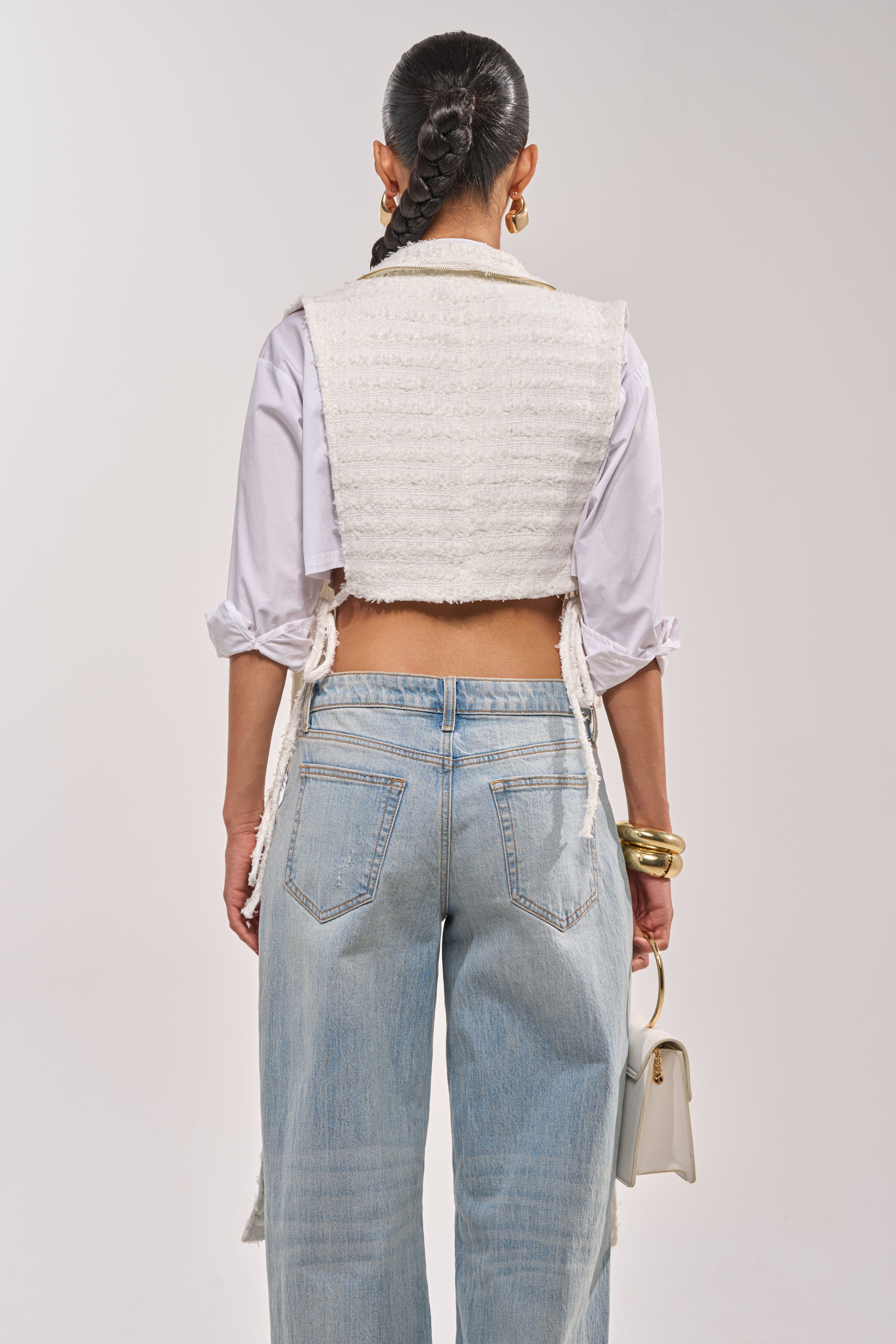 A person with a long braided ponytail is seen from behind wearing the HONEY HONEY SPLIT VEST IN TWEED over a white shirt with rolled sleeves, light blue jeans, gold bangles, and holding a small white handbag. 2