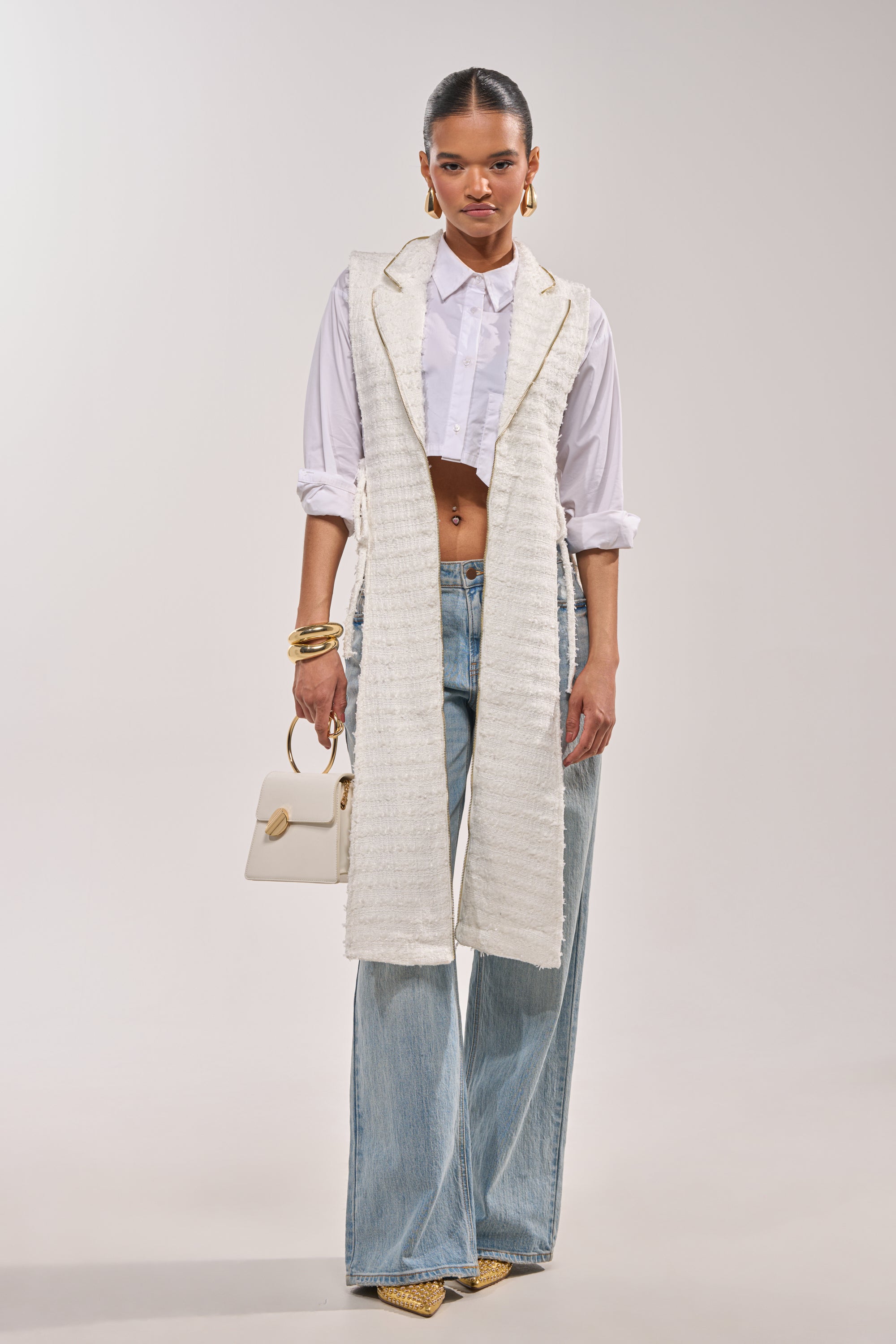 A woman poses against a plain background wearing the HONEY HONEY SPLIT VEST IN TWEED over a white cropped shirt, light blue wide-leg jeans, gold shoes and jewelry, and holding a small white handbag. 3