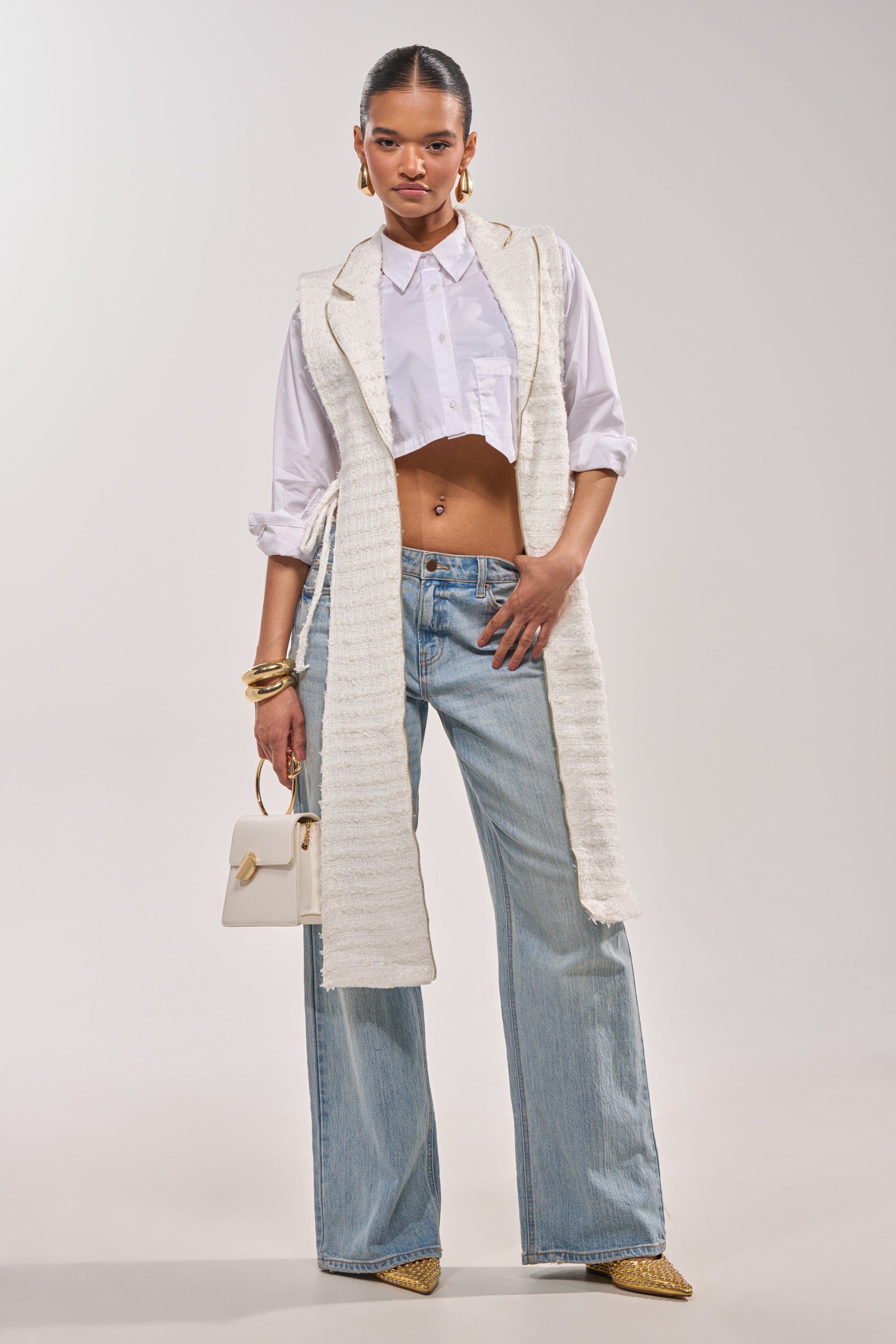 A woman poses confidently in a cropped white button-up, HONEY HONEY SPLIT VEST IN TWEED, light blue wide-leg jeans, pointed yellow shoes, gold accessories, and completes her look with a small white handbag. 4