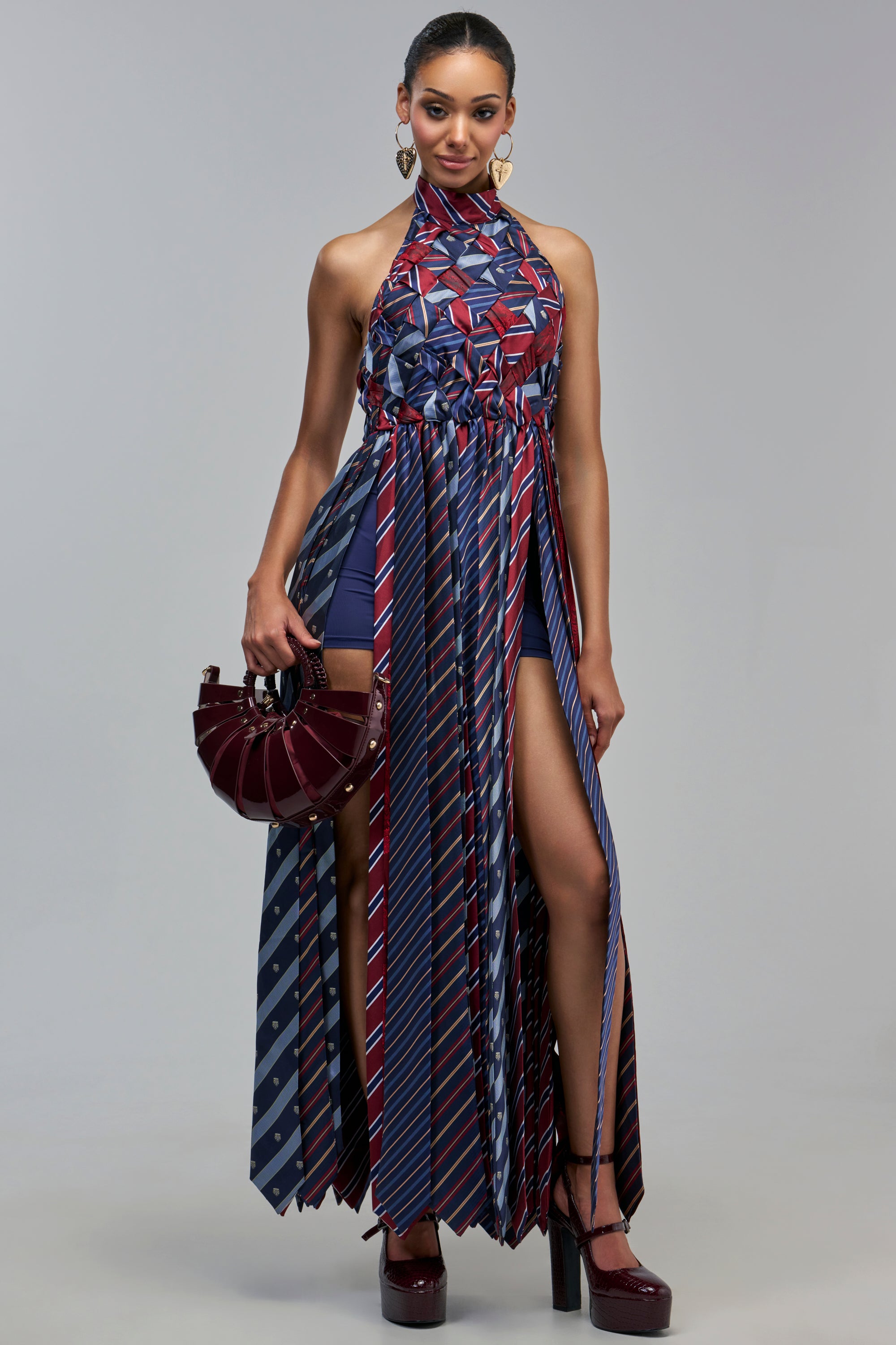 A person wears the SLEEK AUTHORITY HIGH NECK MAXI DRESS, a maximalist piece made of striped and crested neckties with dual high slits over navy shorts, styled with a dark red semi-circular handbag and platform heels.