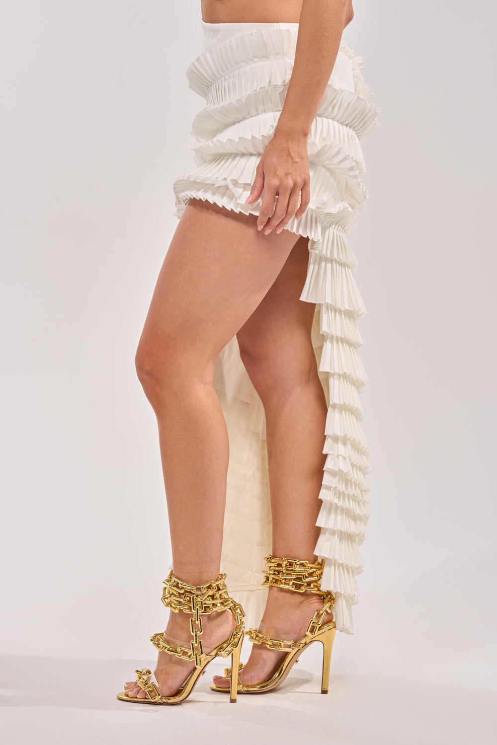 Wearing the DANCE THE NIGHT AWAY RUFFLE SKIRT—a white, high-rise pleated skirt with layered details—paired with gold heeled sandals featuring chunky chain accents, standing against a plain light background. 6