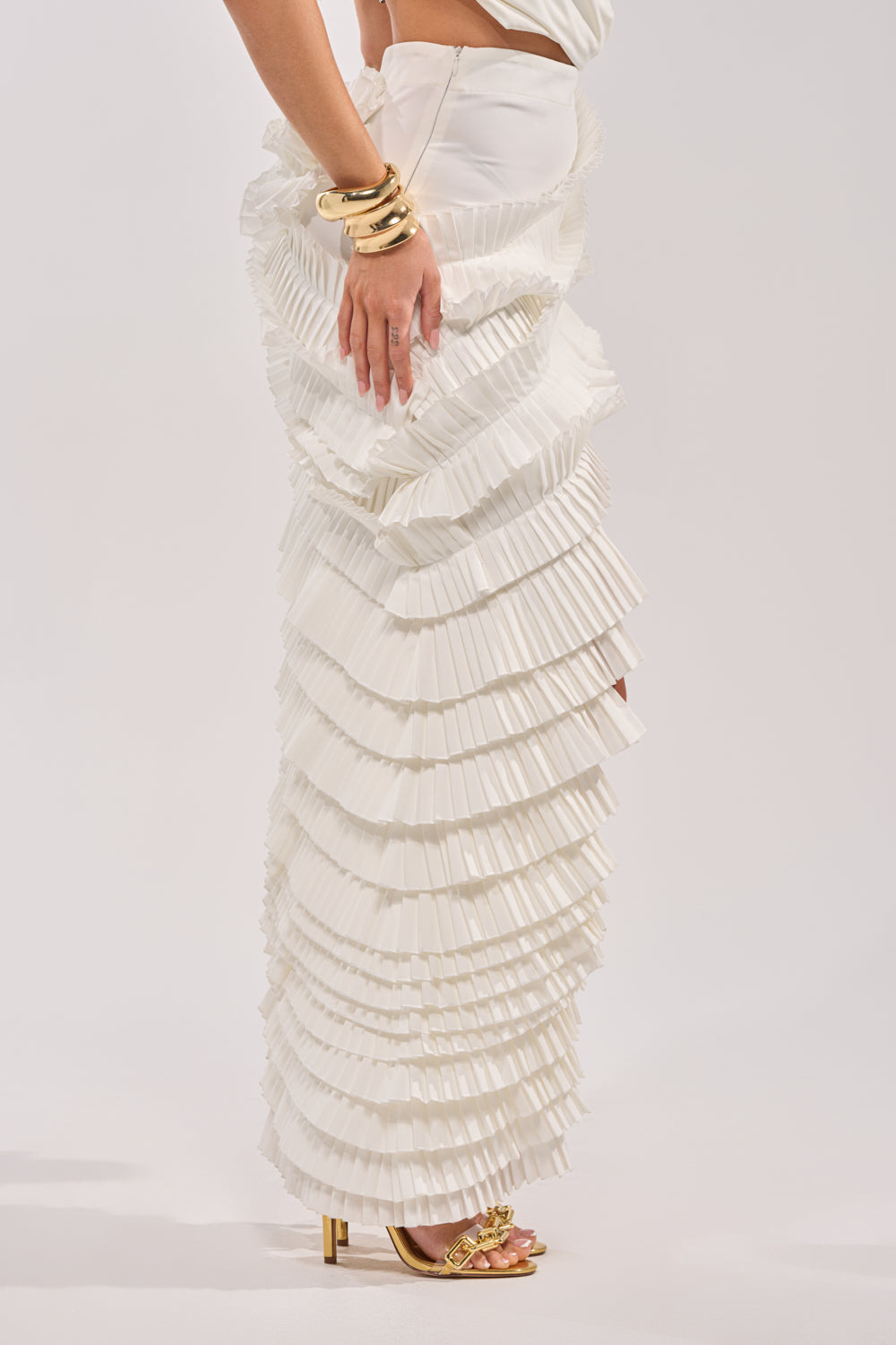 Wearing the DANCE THE NIGHT AWAY RUFFLE SKIRT, a long white pleated skirt with ruffles, paired with gold bracelets and gold high-heeled sandals, a person stands against a plain background. 7