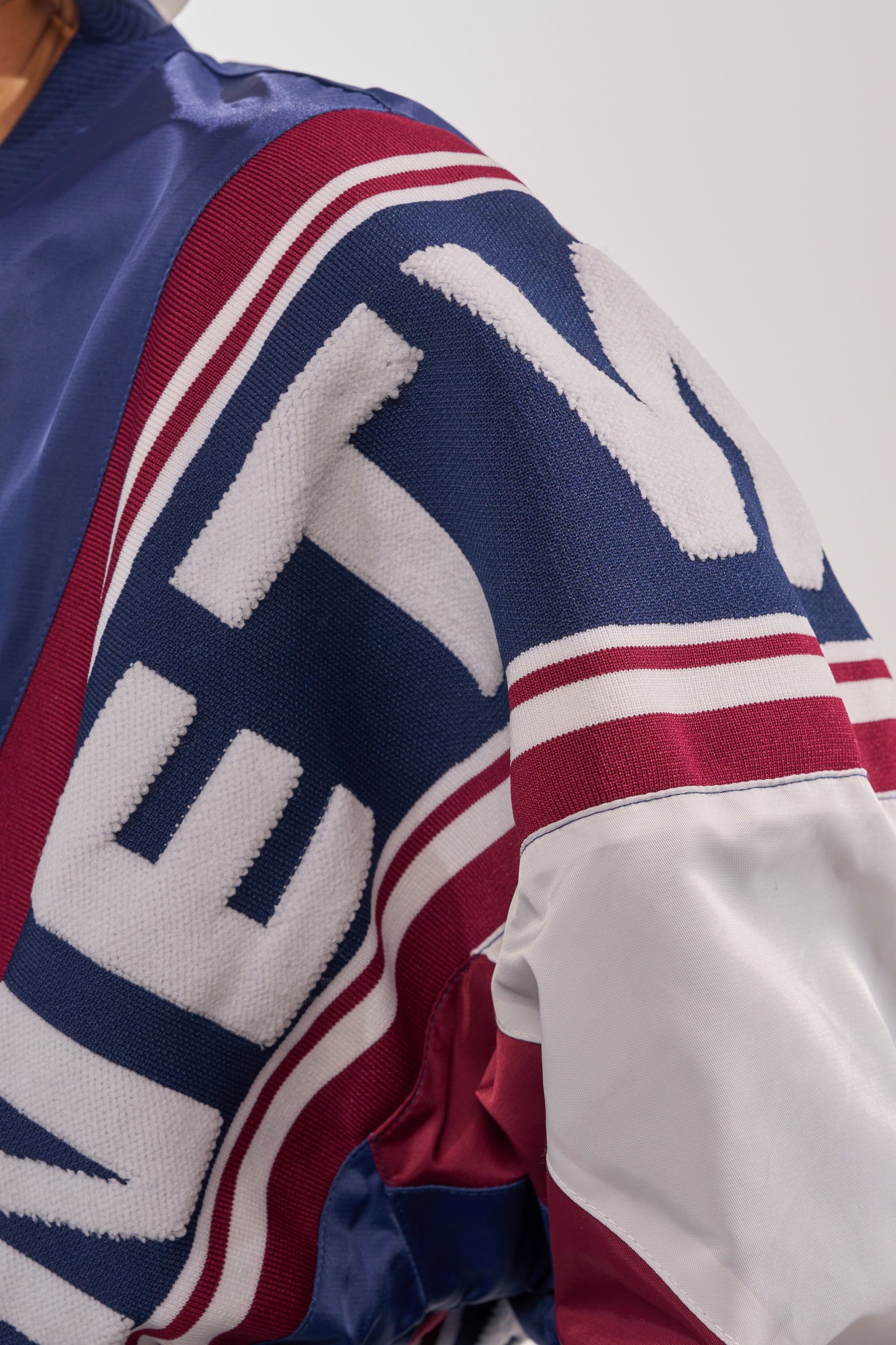 A close-up of the PHOENIX MIXED MEDIA BOMBER IN NAVY shows bold, textured white letters on a navy sleeve with maroon and white stripes, detailed stitching, and a hint of a white section near the elbow. 9