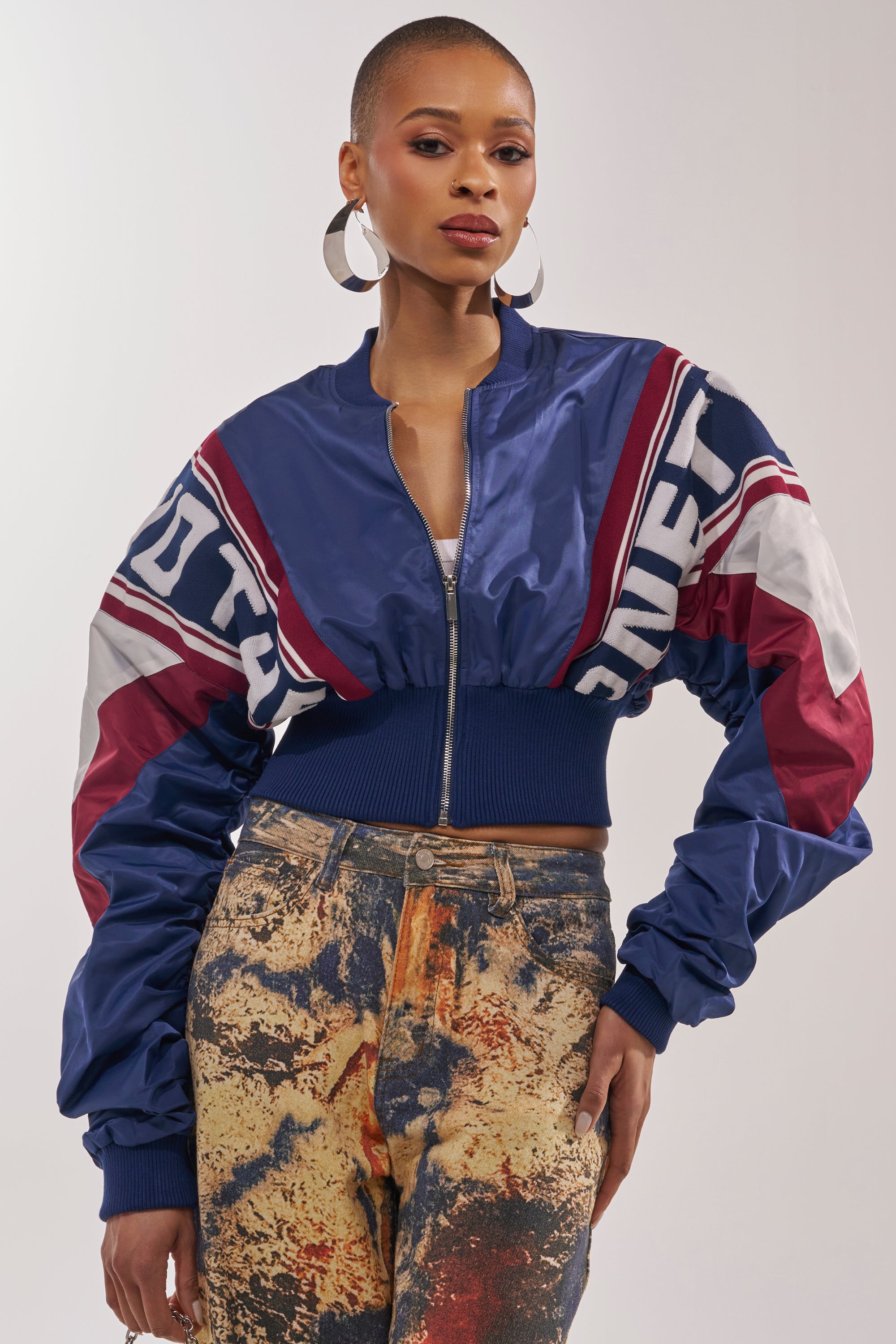 A woman with short hair wears large hoop earrings, high-waisted abstract print jeans, and the PHOENIX MIXED MEDIA BOMBER IN NAVY—a cropped varsity jacket with bold lettering and ruched sleeves in blue, white, and maroon. 2