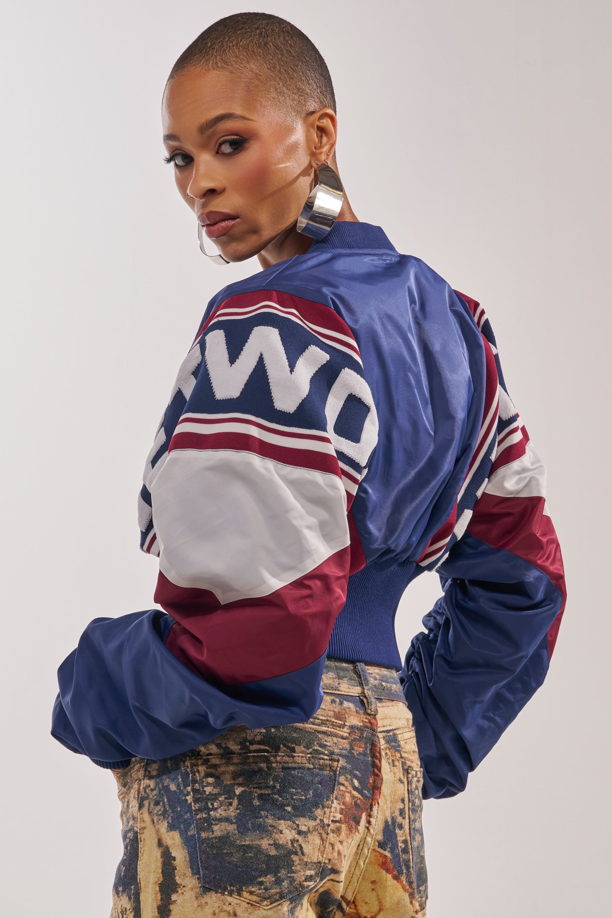 A woman with a shaved head wears large hoop earrings, high-waisted acid-wash jeans, and the PHOENIX MIXED MEDIA BOMBER IN NAVY. She looks over her shoulder against a plain background. 4