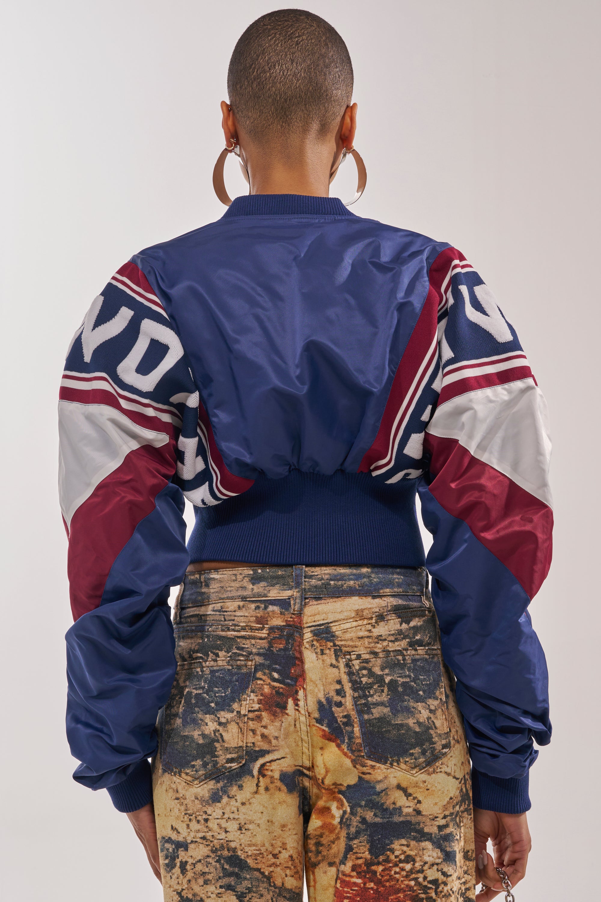 A person with a shaved head and large hoop earrings wears the PHOENIX MIXED MEDIA BOMBER IN NAVY, featuring bold patterns and ruched sleeves, paired with high-waisted, abstract-print jeans. The photo is taken from behind. 5