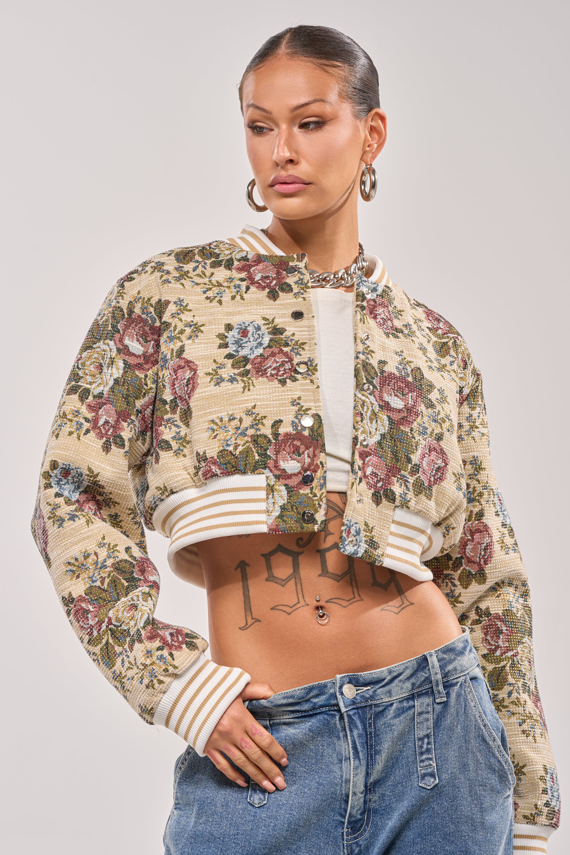 A woman models the NOT YOUR AVERAGE TAPESTRY BOMBER over a white crop top and blue jeans, accessorized with large hoop earrings. Her slicked-back hair and visible torso tattoos complete the bold, stylish look as she poses with one hand in her pocket. 0