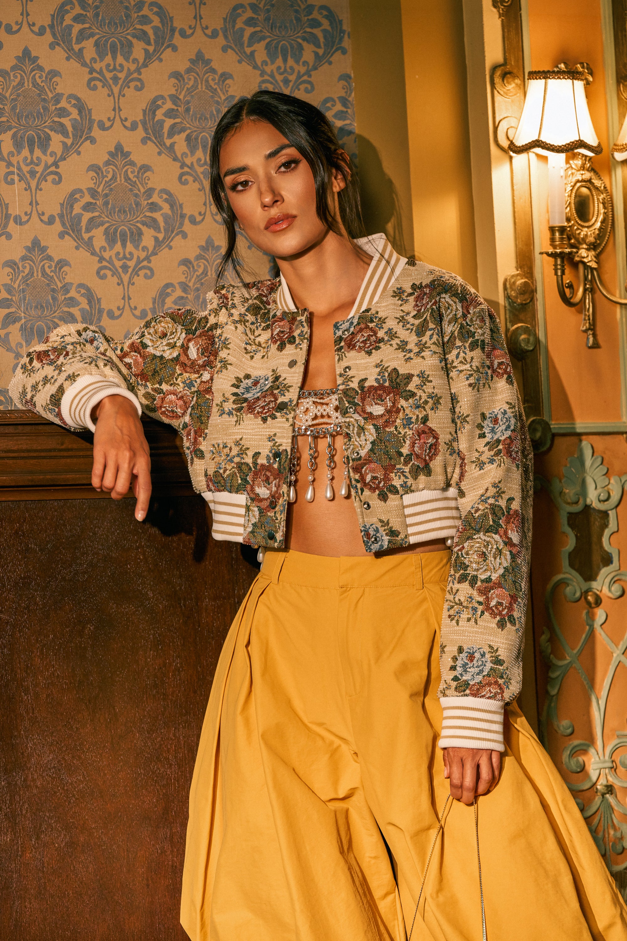 A woman wearing the NOT YOUR AVERAGE TAPESTRY BOMBER, paired with a beaded crop top and wide yellow pants, stands indoors against ornate wallpaper, resting one arm on a wooden ledge near a vintage-style lamp.