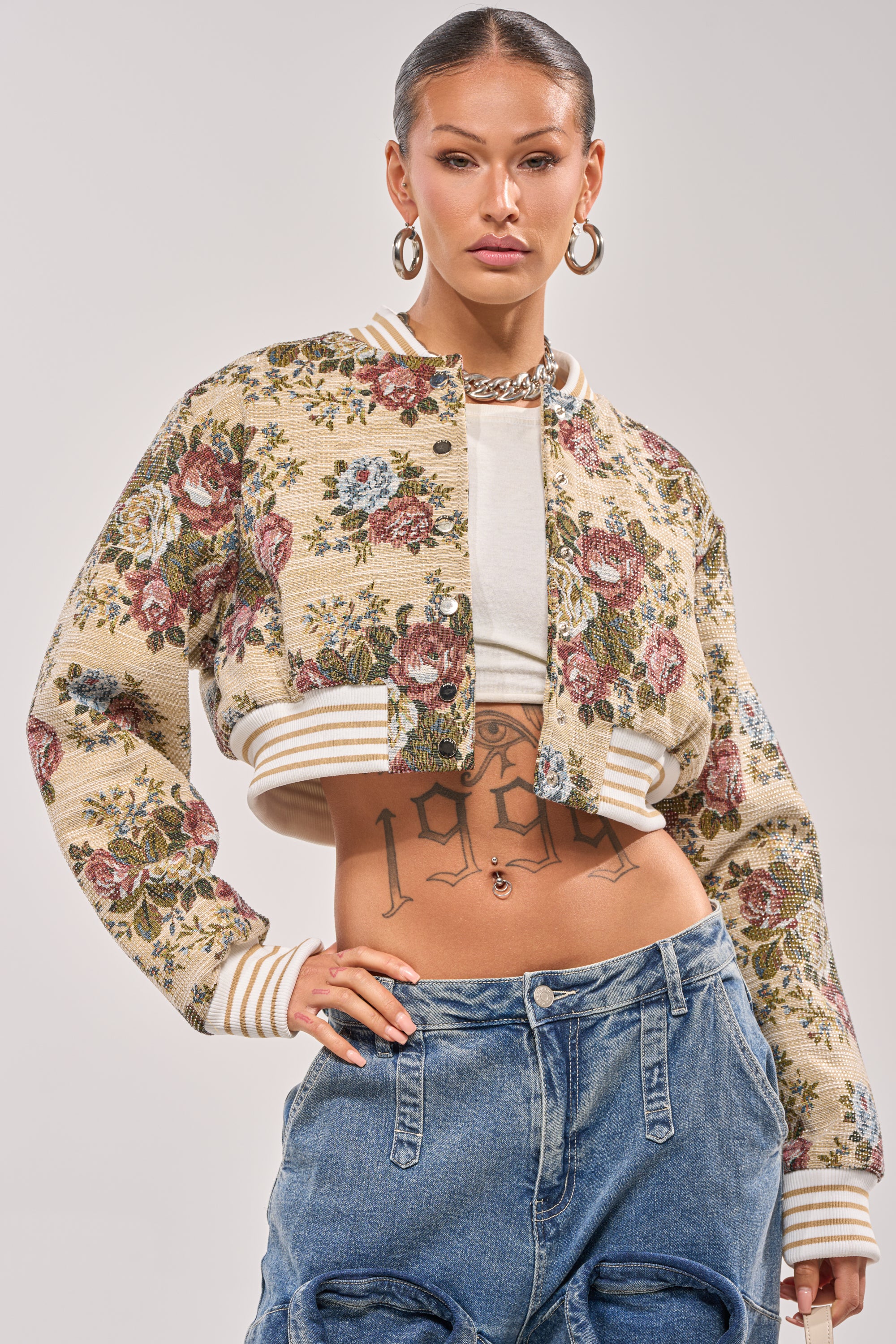 A woman wears the NOT YOUR AVERAGE TAPESTRY BOMBER over a white top, revealing a torso tattoo. She pairs it with blue jeans and hoop earrings, finishing the look with slicked-back hair. 1