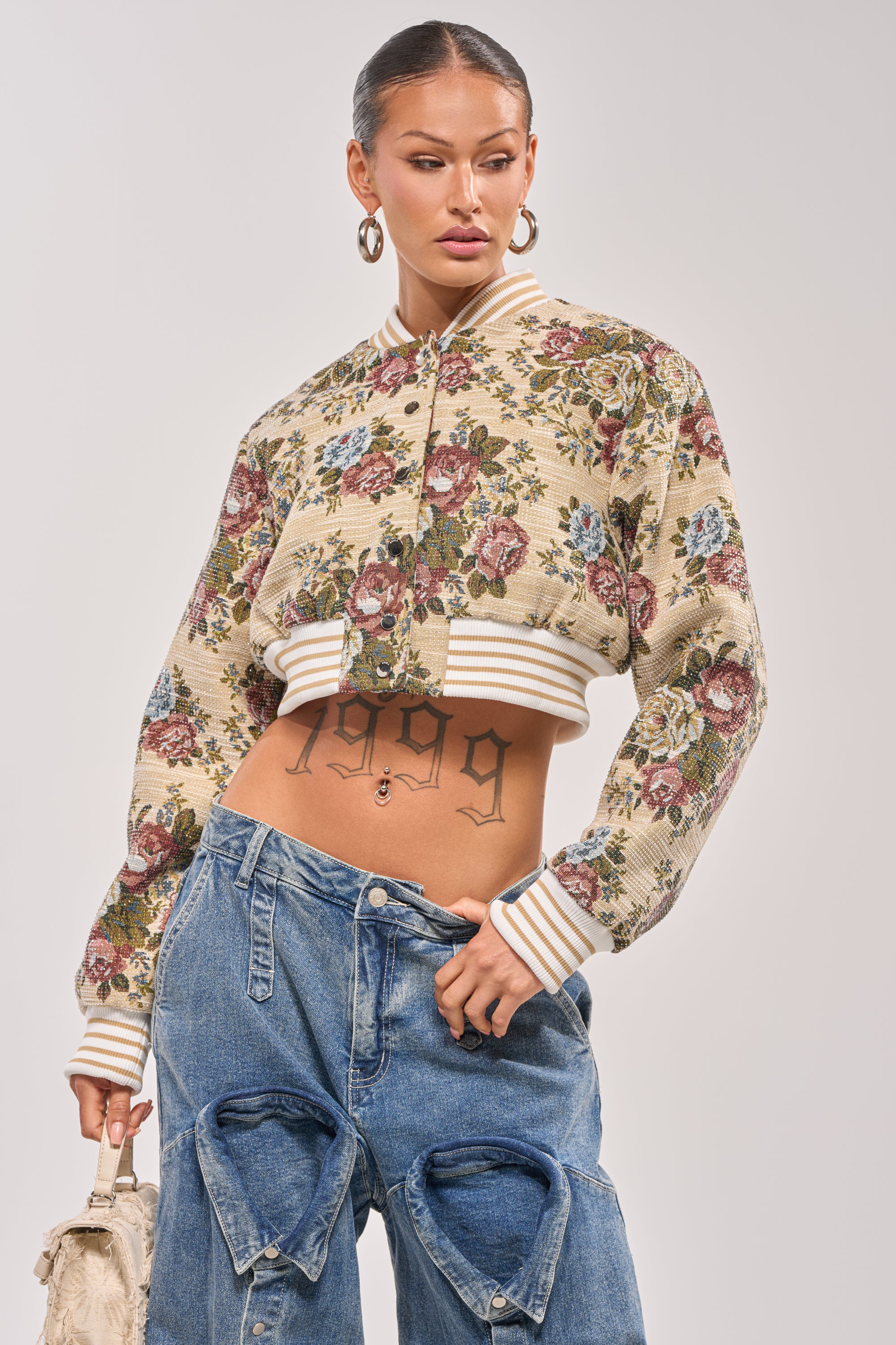 A woman wearing the NOT YOUR AVERAGE TAPESTRY BOMBER, loose ripped blue jeans, and hoop earrings poses confidently with a beige textured handbag. Her "1999" stomach tattoo is visible. 2