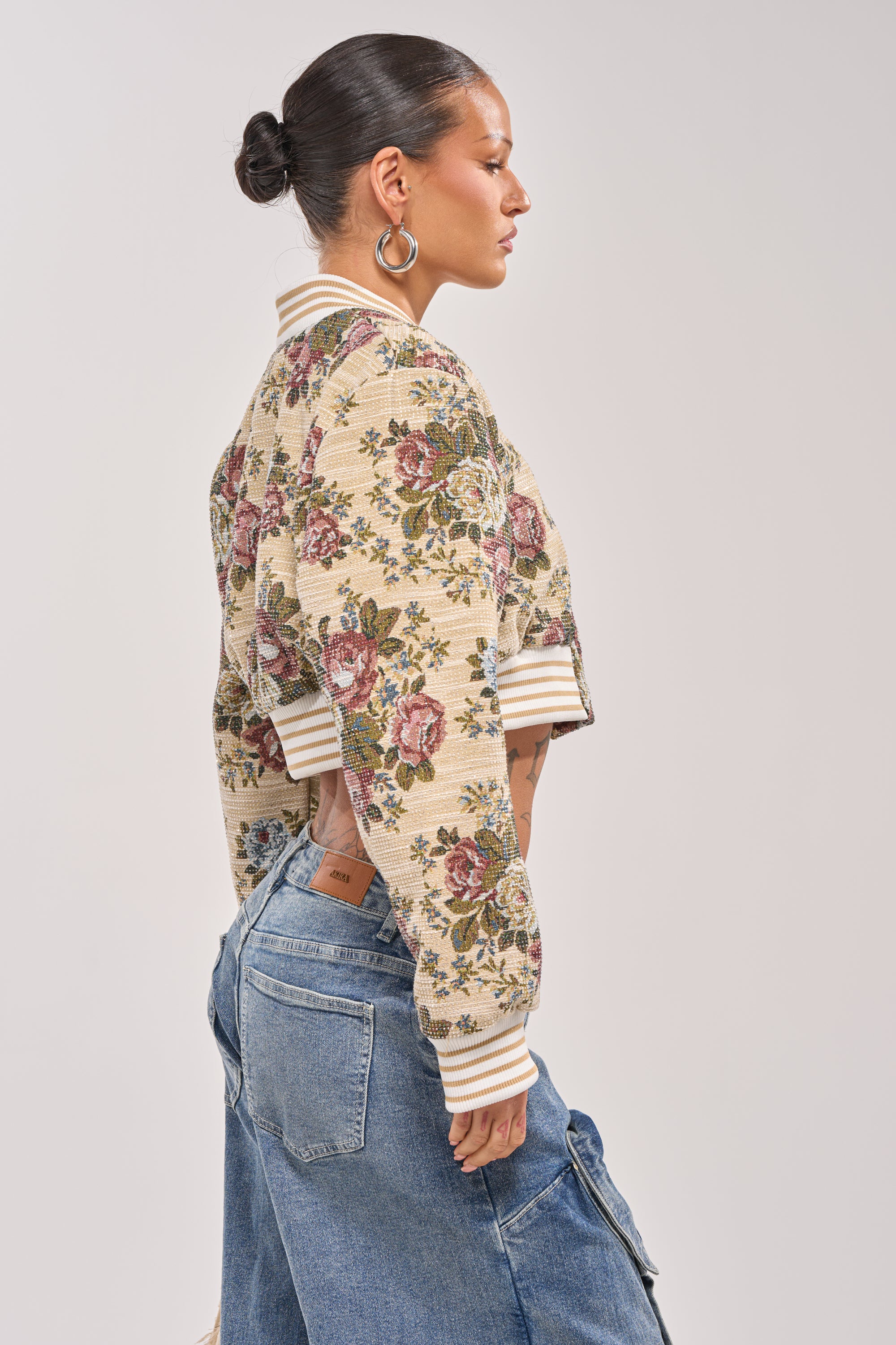 A woman stands in profile wearing the NOT YOUR AVERAGE TAPESTRY BOMBER—a beige floral jacket with striped cuffs and collar—paired with patchwork blue jeans and large hoop earrings. Her neatly tied-back hair highlights the standout bomber. 3