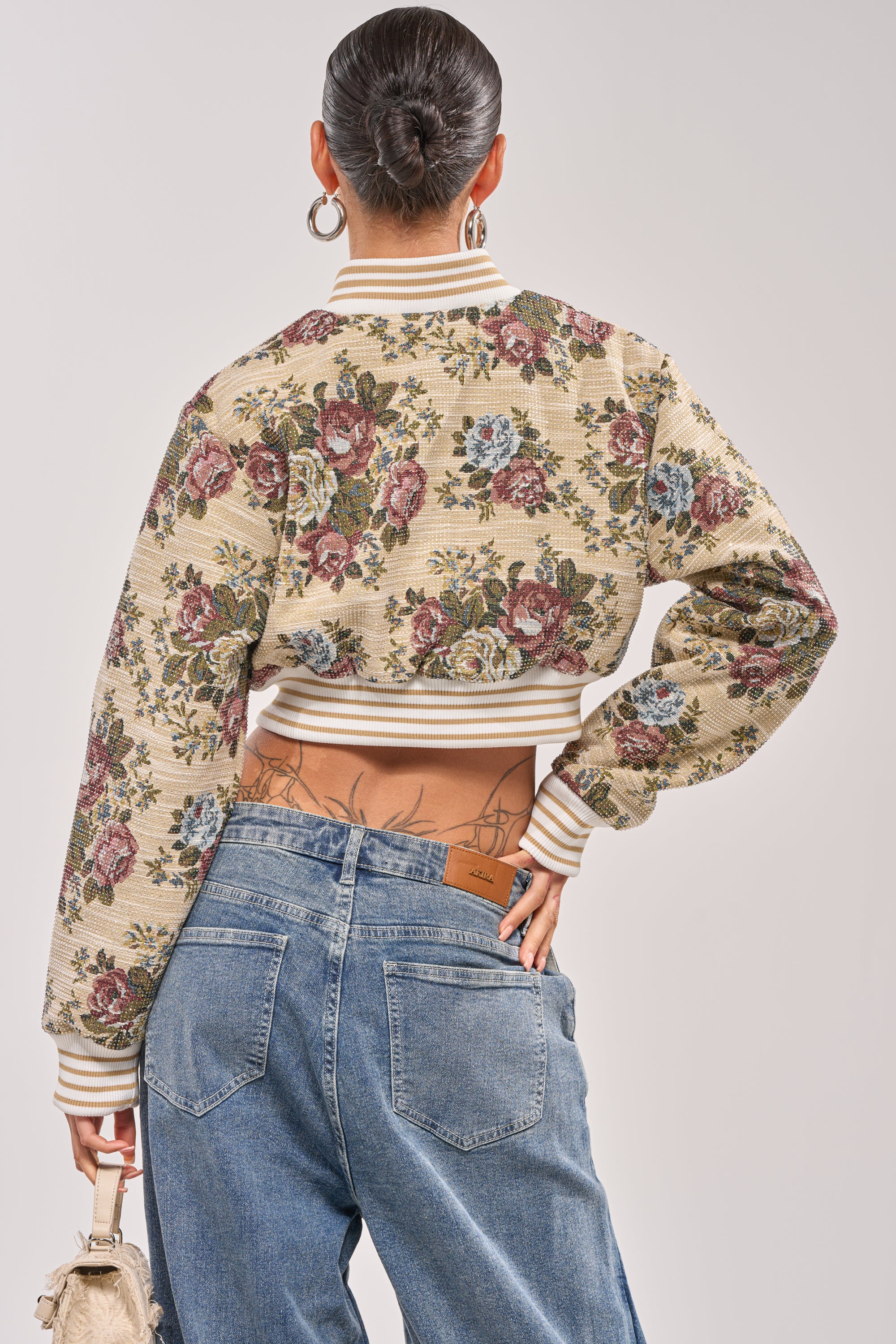A person with a bun hairstyle wears the NOT YOUR AVERAGE TAPESTRY BOMBER and loose blue jeans, standing with their back to the camera and holding a small bag, revealing a tattoo on their lower back. 4