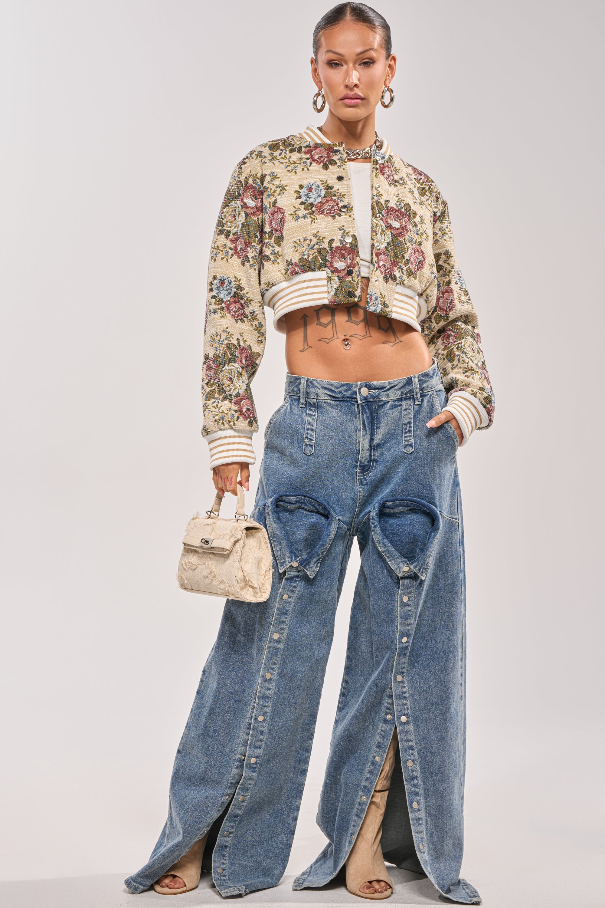 A woman stands confidently in the NOT YOUR AVERAGE TAPESTRY BOMBER, wide-leg jeans with heart cutouts, beige open-toe heels, large hoop earrings, and a small beige handbag. 5