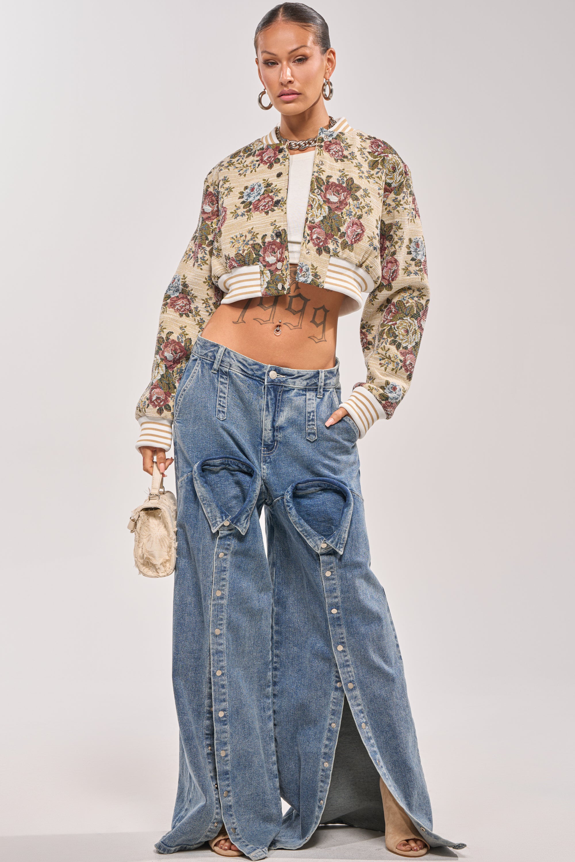 A woman models the NOT YOUR AVERAGE TAPESTRY BOMBER, pairing it with wide-leg blue jeans featuring large heart cutouts. She accessorizes with a small beige bag, hoop earrings, and has her hair tied back. 6