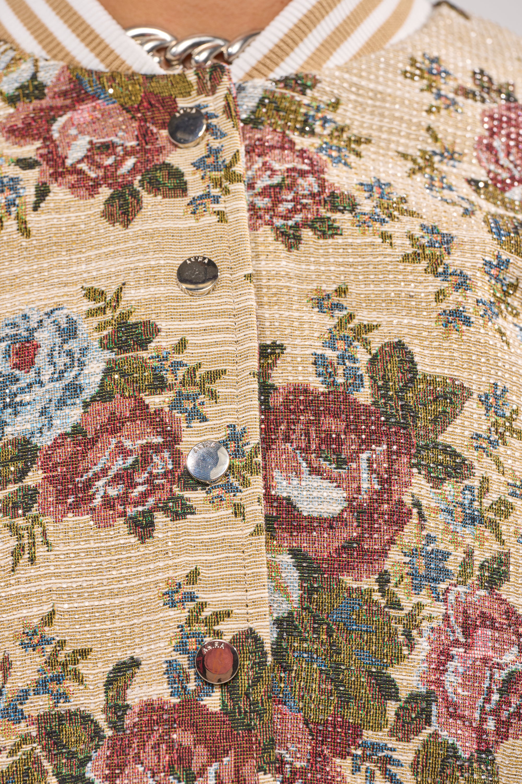 Close-up of the NOT YOUR AVERAGE TAPESTRY BOMBER in beige with a textured floral pattern in red, blue, and green, metallic snap buttons, and a striped beige and white collar. 7