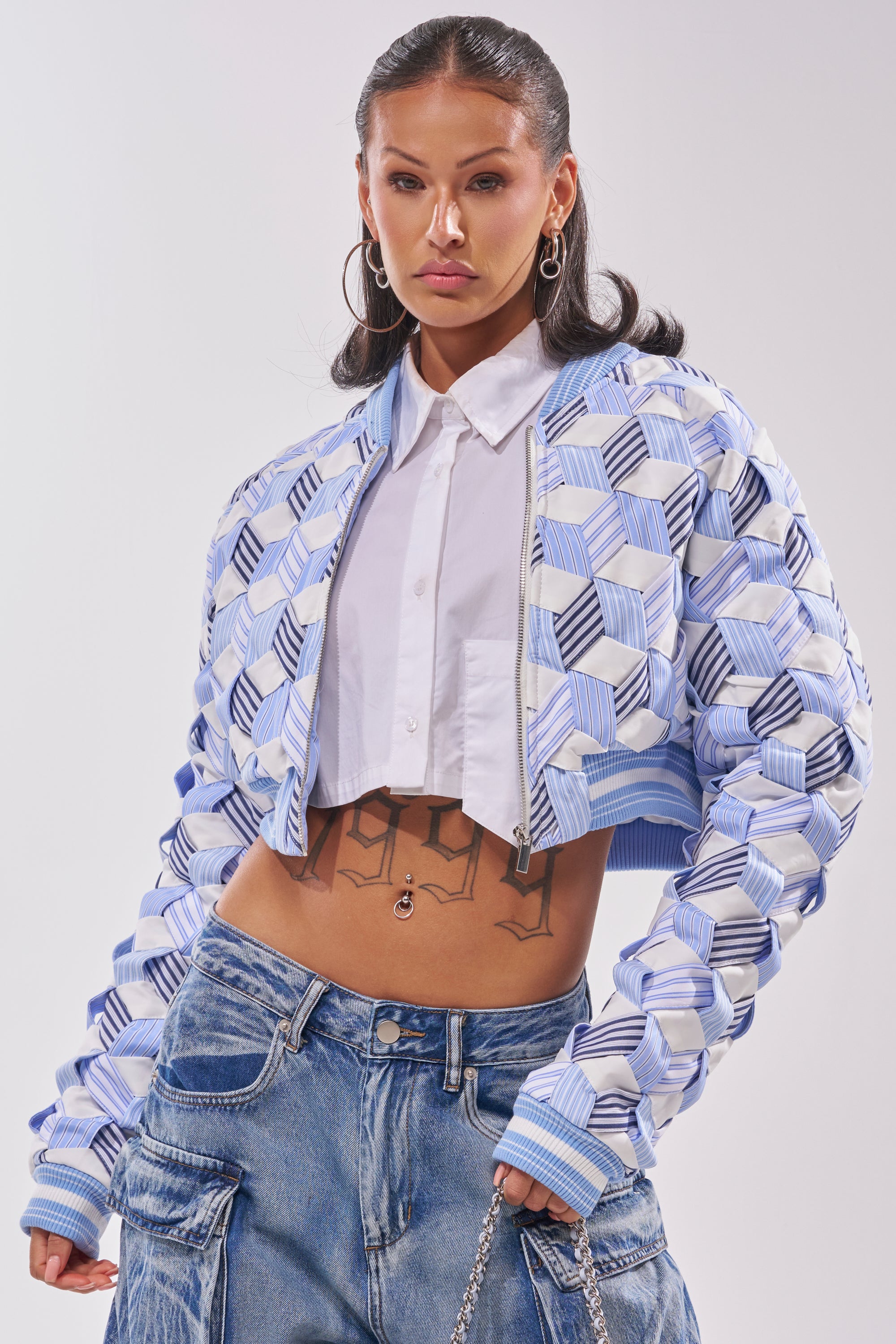 A woman with long dark hair wears the BOP AND WEAVE POPLIN BOMBER over a white cropped shirt and loose blue jeans, accessorized with large hoop earrings and showing tattoos on her torso. 0