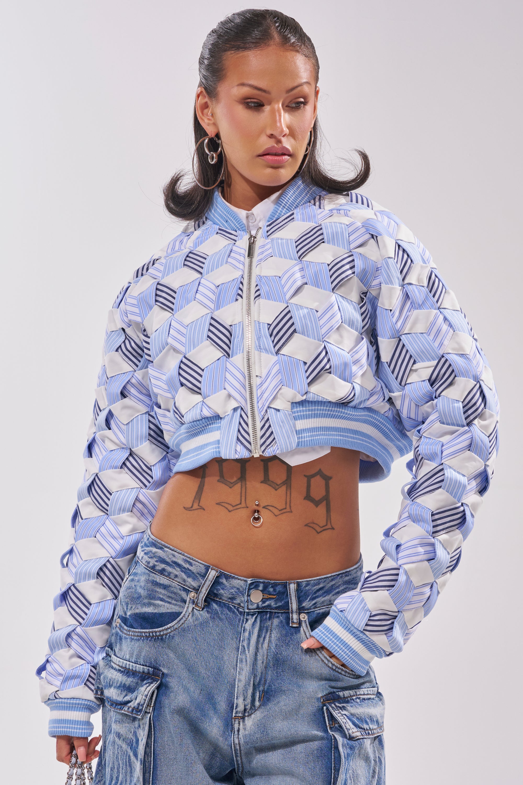 A woman with hoop earrings and slicked-back hair wears the BOP AND WEAVE POPLIN BOMBER—a blue and white geometric cropped jacket—with baggy jeans, showing a “1999” tattoo on her midriff as she looks to the side against a plain backdrop. 2