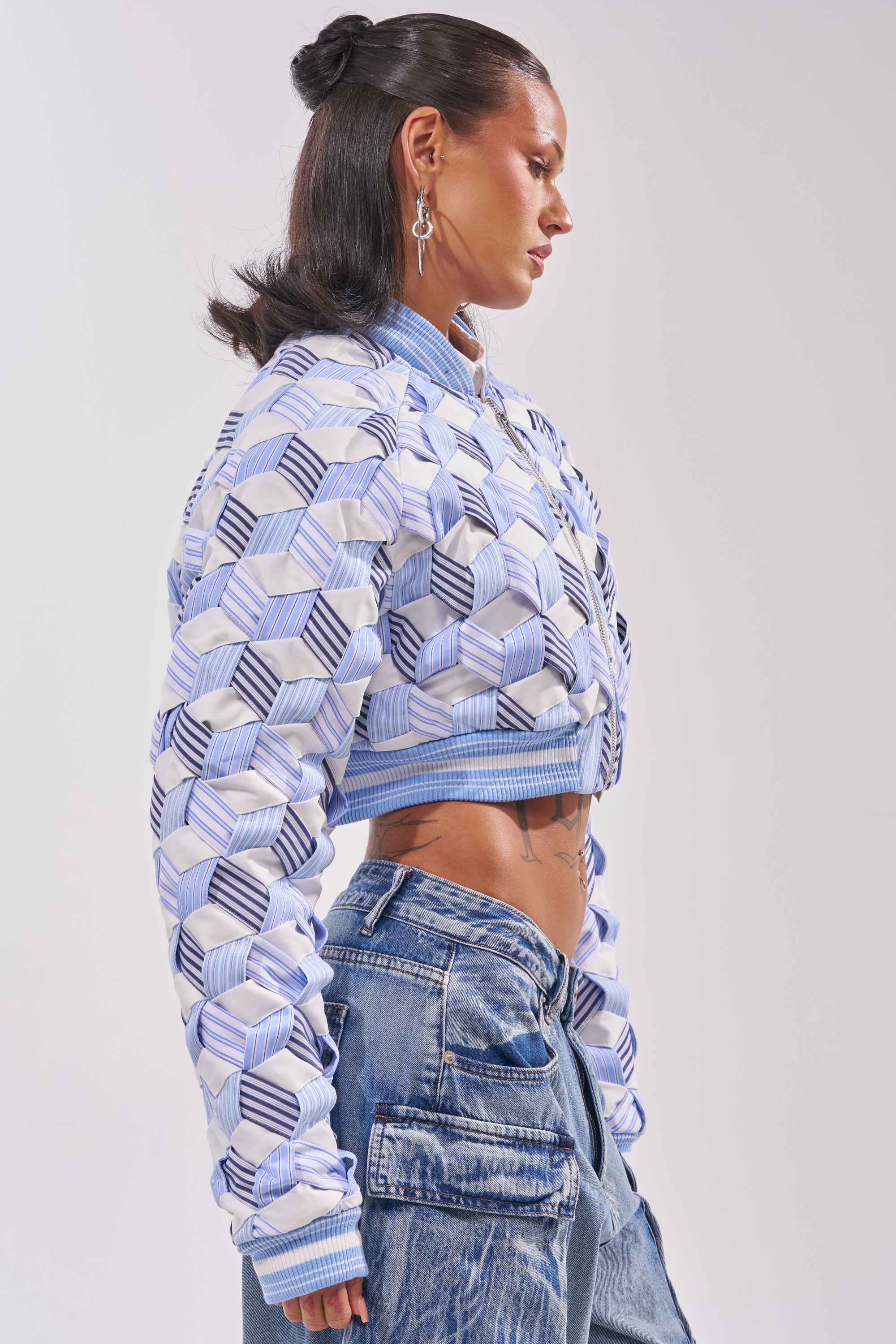 A person in profile wears the BOP AND WEAVE POPLIN BOMBER, featuring a blue and white geometric pattern, paired with loose, low-rise blue jeans that reveal their midriff. Their hair is sleek and half-up, complemented by dangling earrings. 3