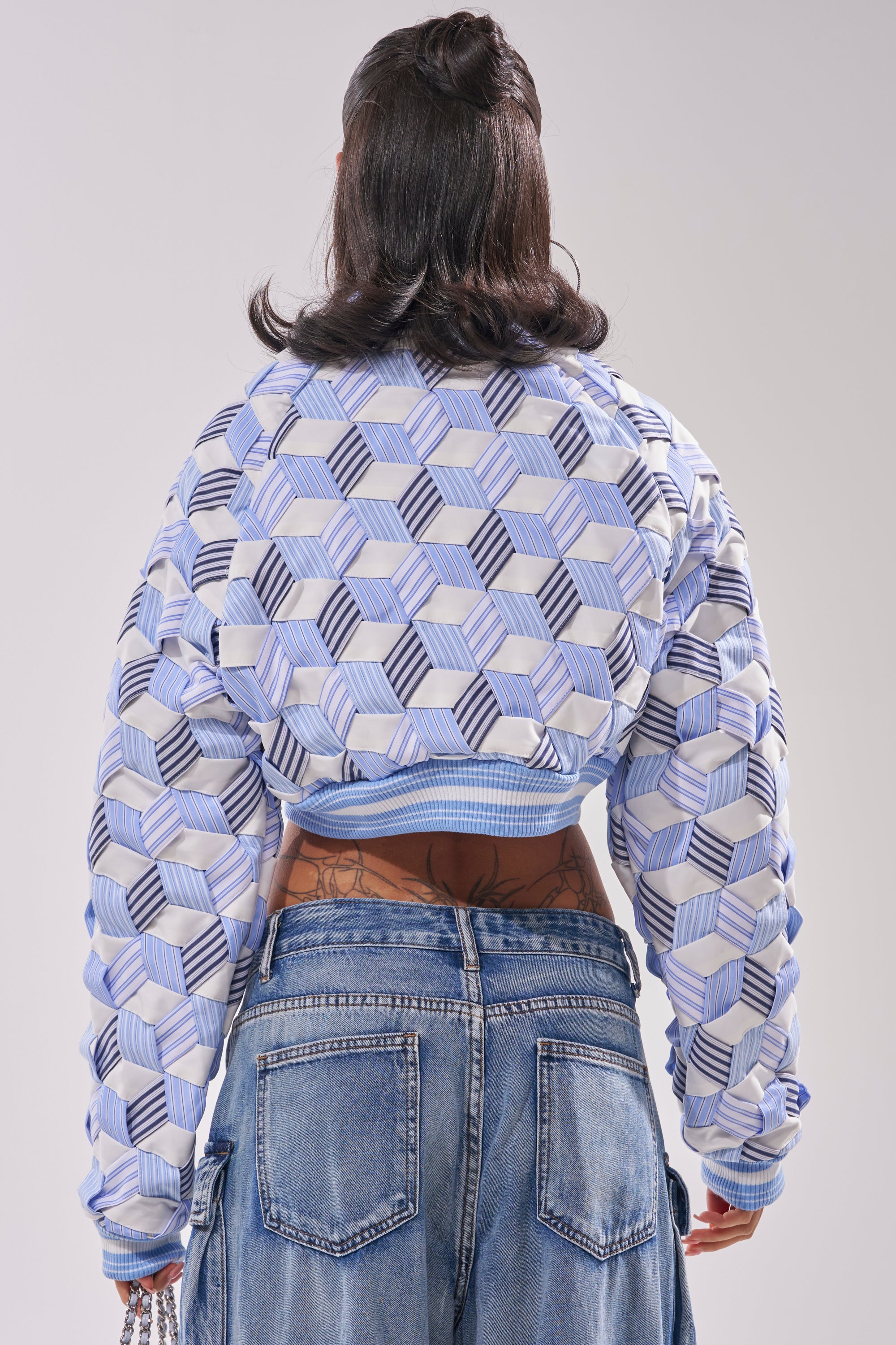 A person stands facing away, wearing the BOP AND WEAVE POPLIN BOMBER—blue and white striped and cropped—with loose, high-waisted blue jeans. Their hair is in loose waves, showing tattoos on their lower back. 4
