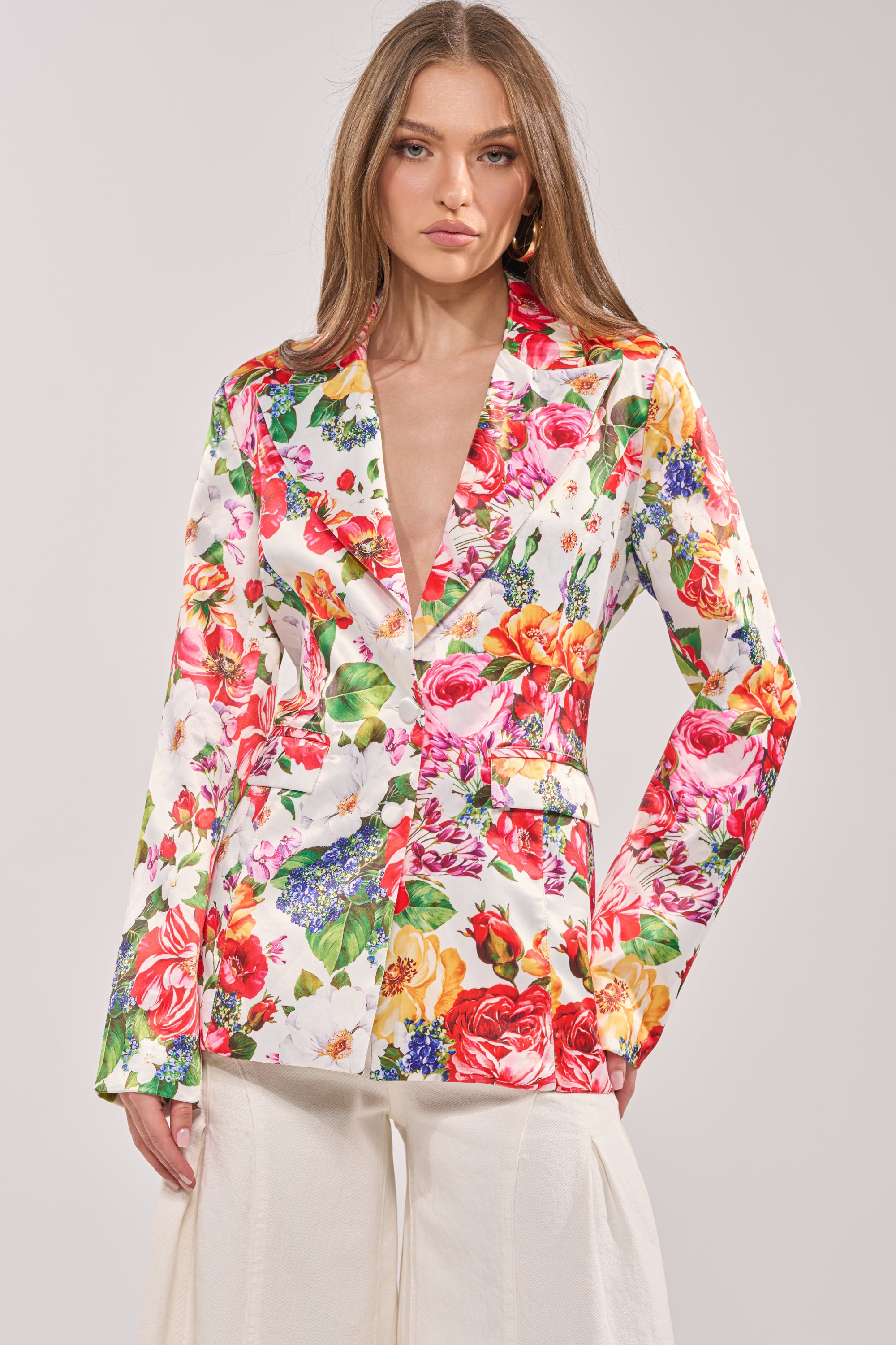 A woman stands confidently against a plain background, wearing the ALESSANDRA SATIN BLAZER featuring a colorful floral pattern, deep neckline, and paired with light wide-leg pants. 0