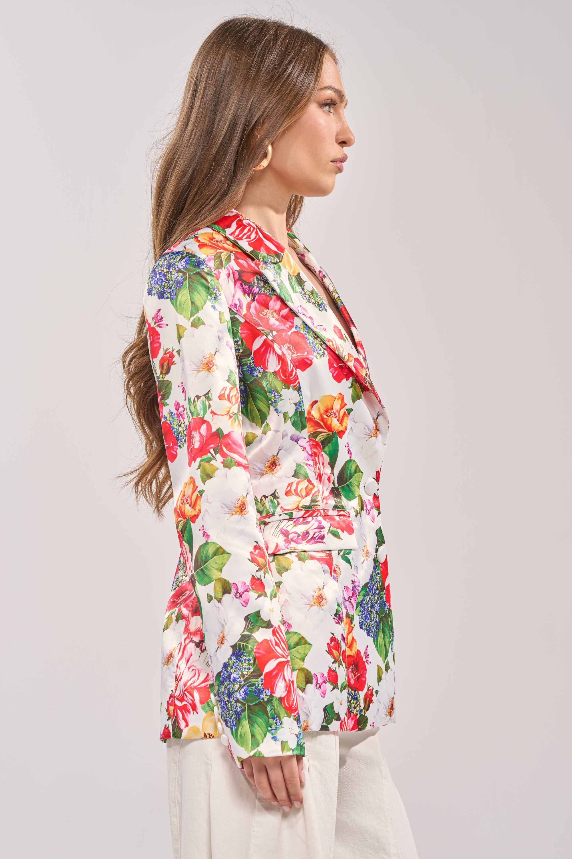 A woman with long brown hair stands in profile against a plain light background, wearing the colorful ALESSANDRA SATIN BLAZER and white pants. 2